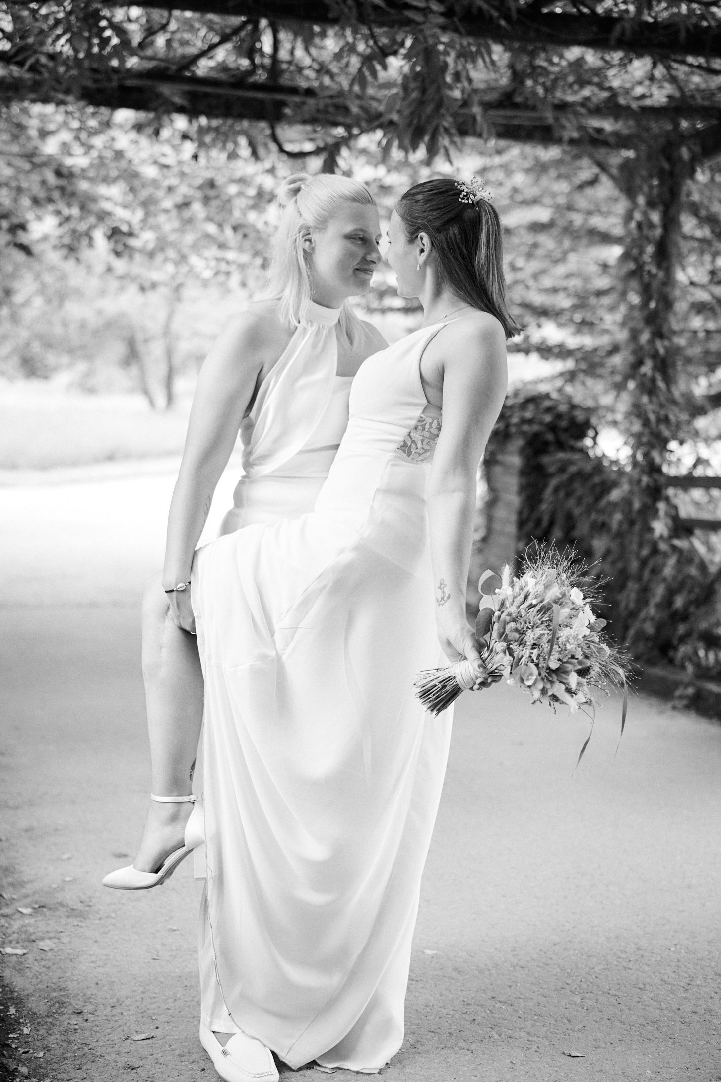 Two brides in white gowns share a romantic moment under a garden pergola, one holding a bouquet, in black and white.