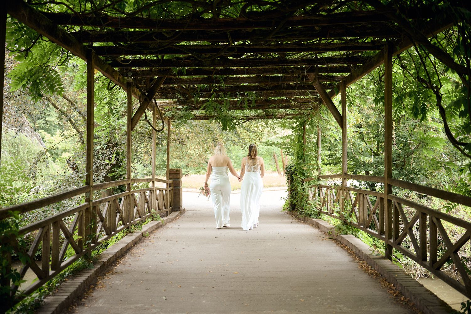 Two brides in white dresses walk hand-in-hand through a lush garden pergola bridge on their wedding day.