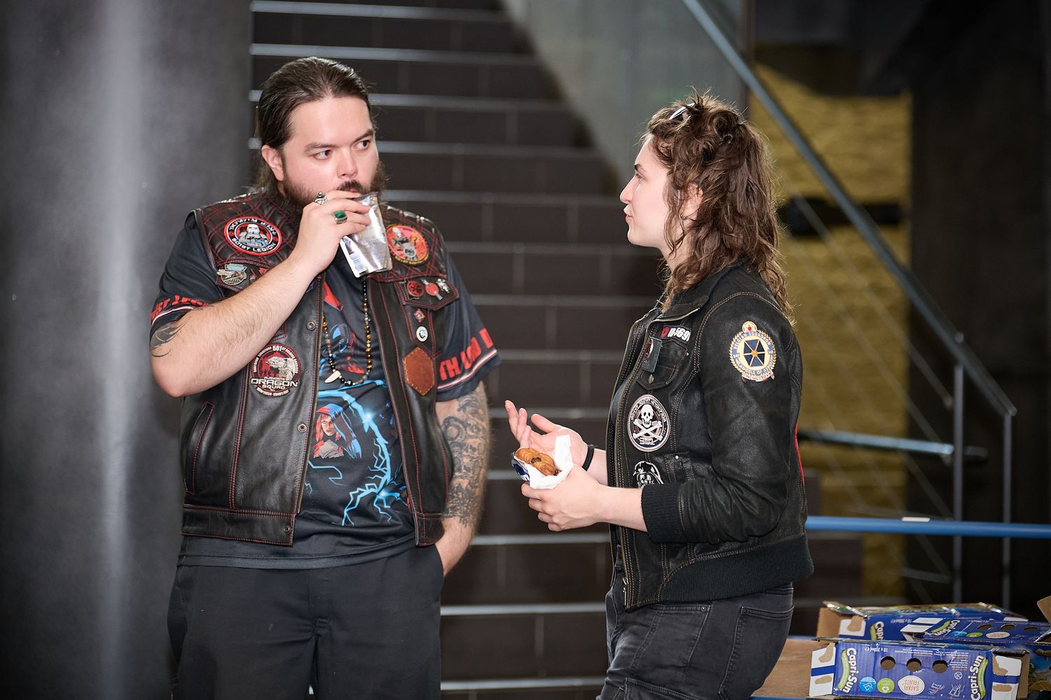 Two men in biker jackets with patches stand talking near stairs, one holding a microphone.
