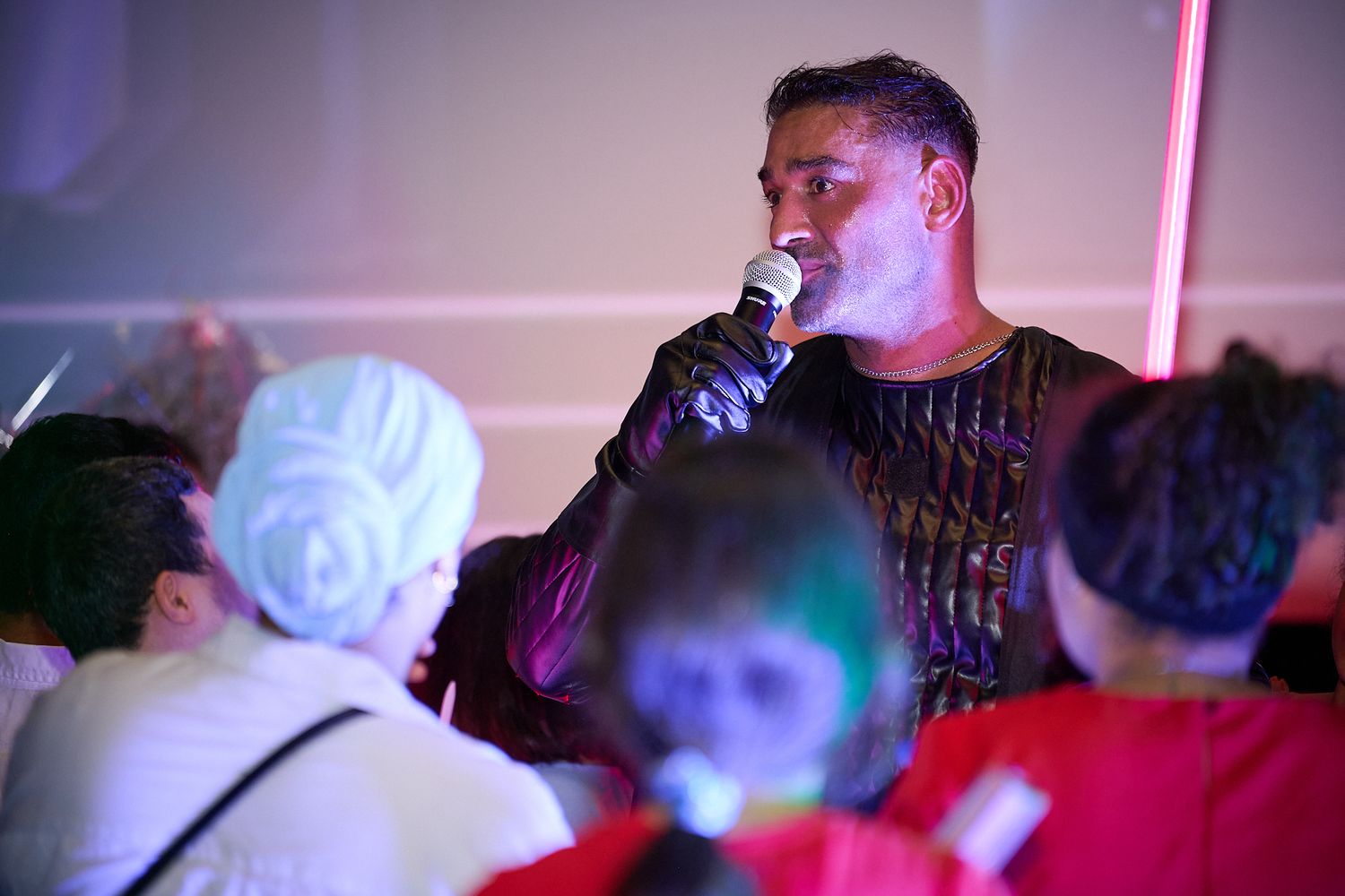 Man speaking into microphone at a colorful indoor event with audience members seated in foreground.