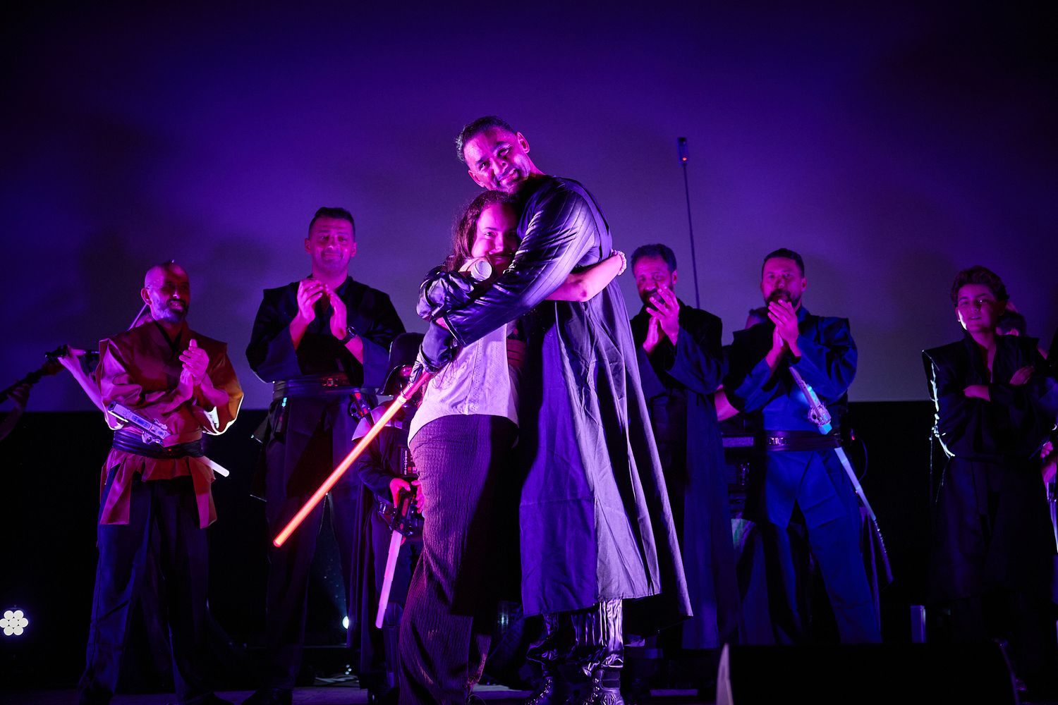 Two performers embrace on stage holding a lightsaber under purple lights with musicians applauding behind them.
