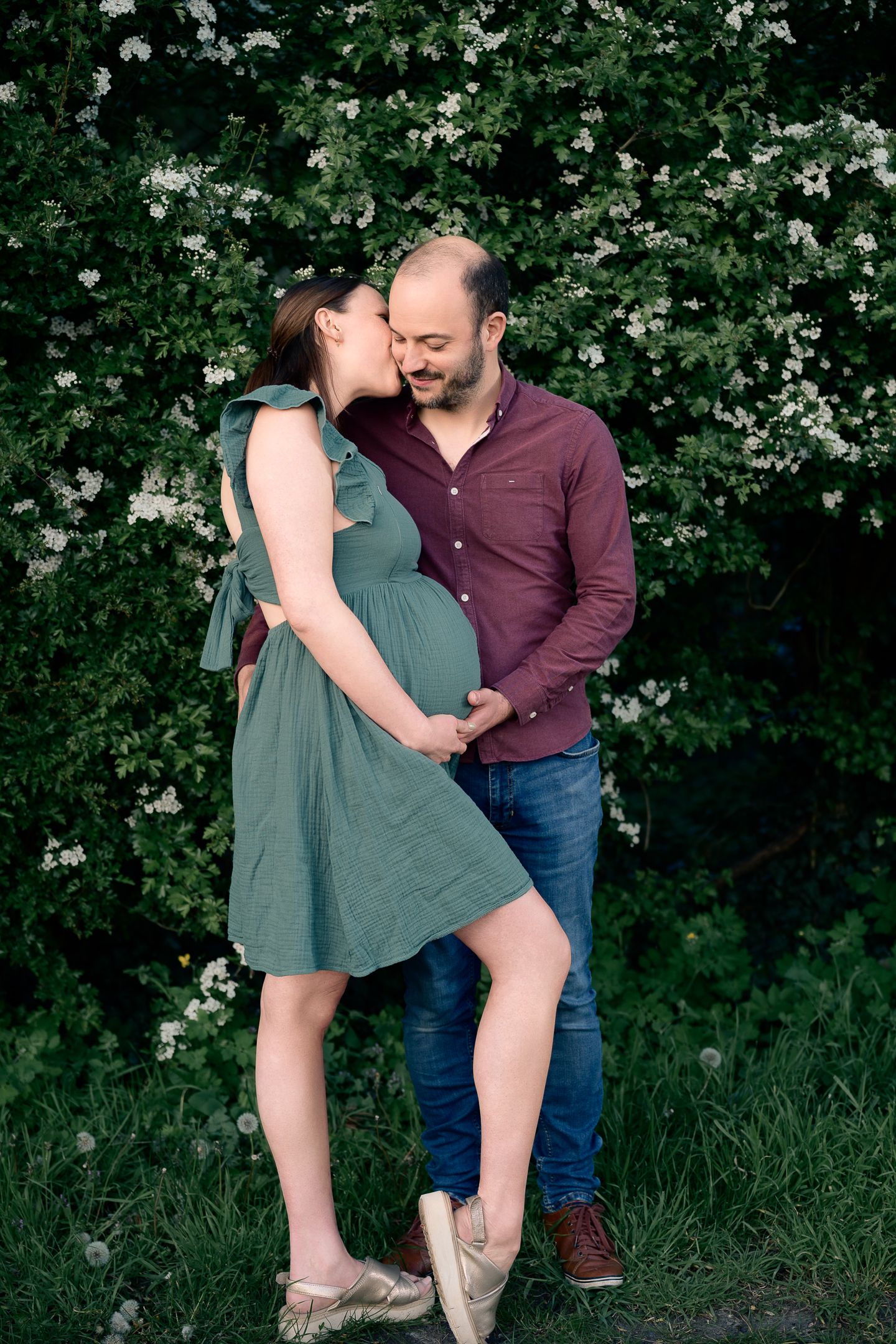 Couple embracing outdoors in front of flowering bushes, pregnant woman in green dress with partner in burgundy shirt.