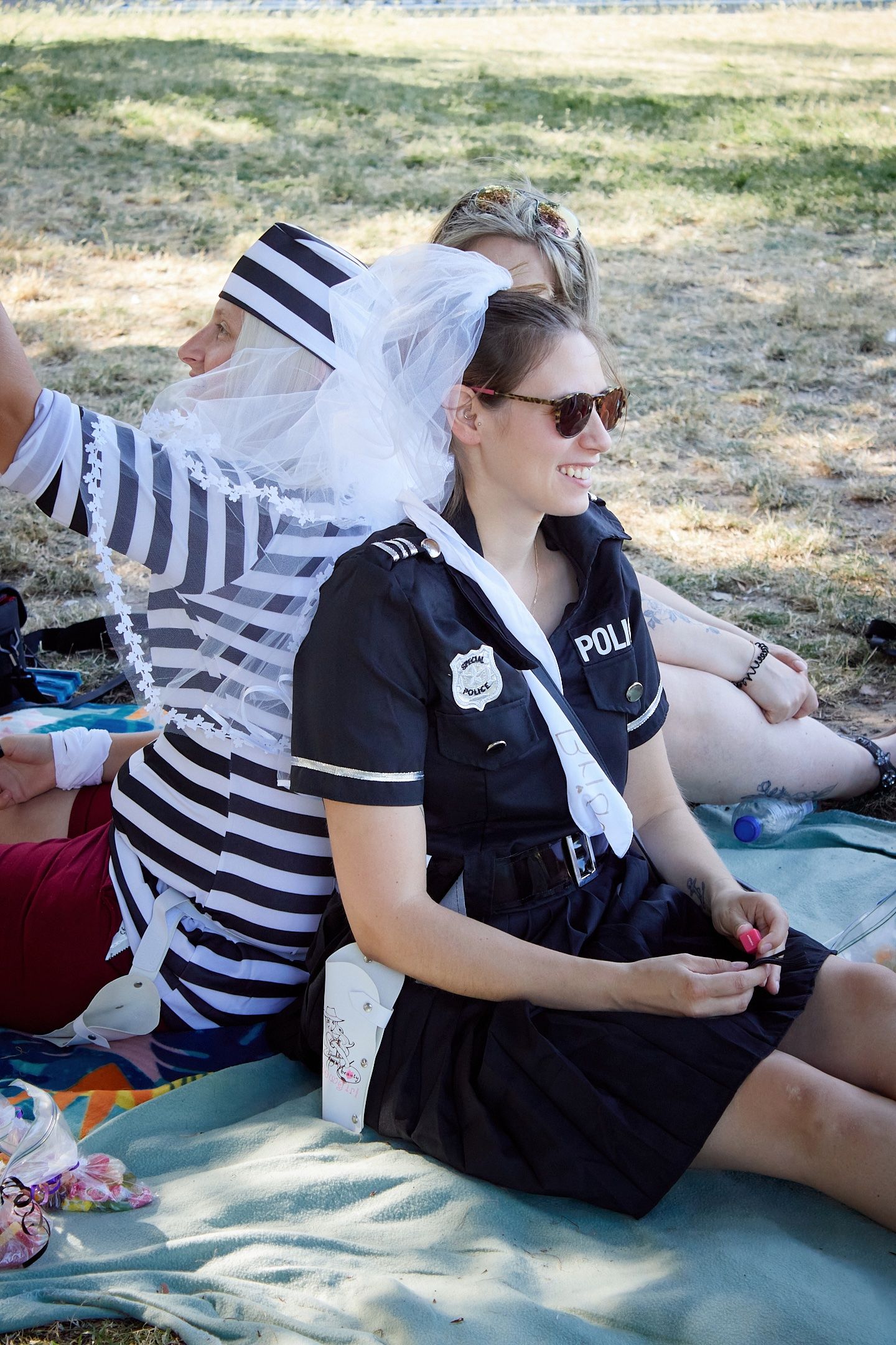 Woman in police costume with veil smiles at outdoor bachelorette party, sitting on blanket with friend in prisoner outfit.