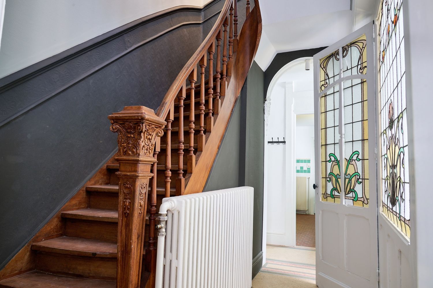 Elegant Victorian hallway featuring ornate wooden staircase, stained glass front door, and white radiator.