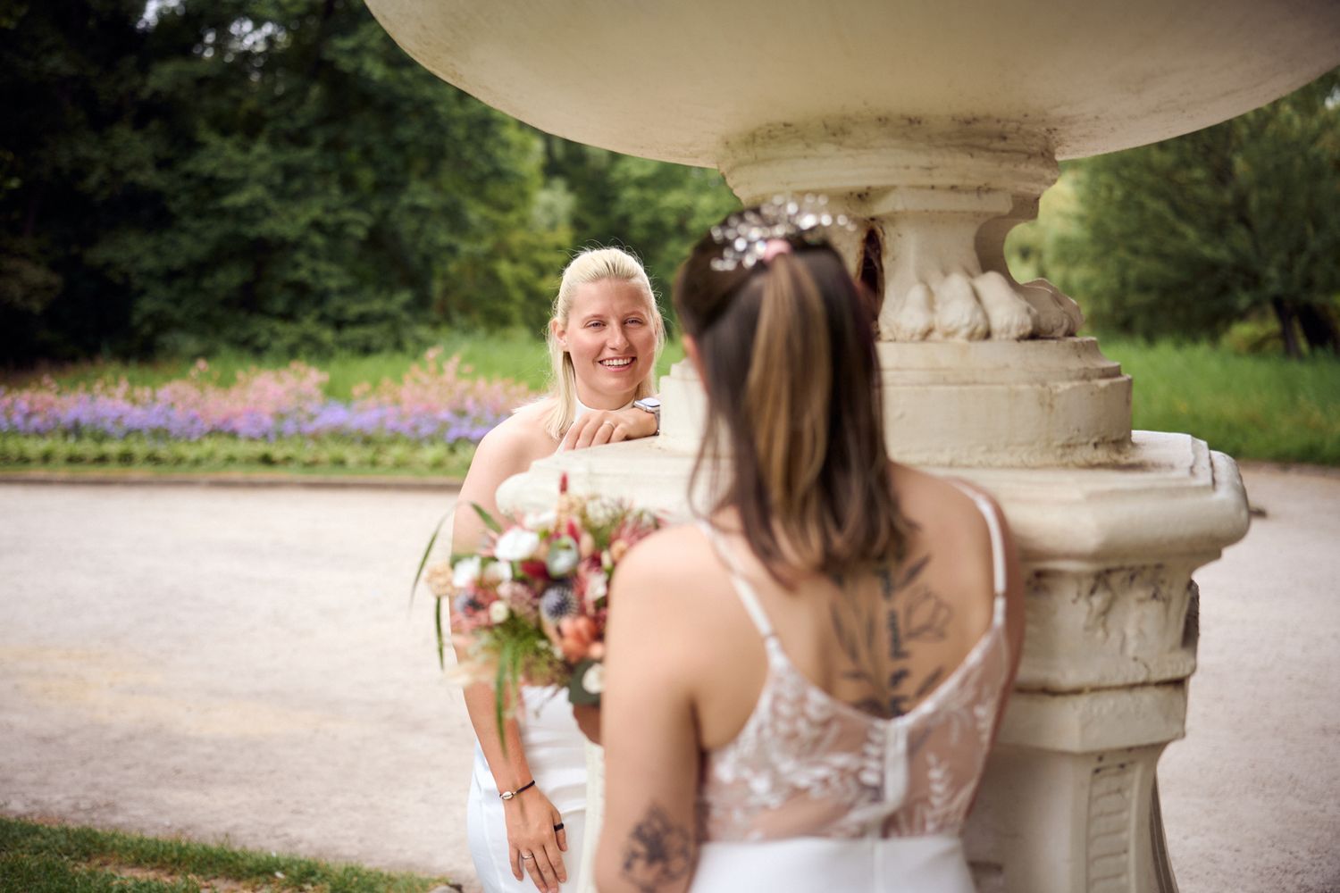 Two brides in white dresses share a joyful moment near a stone fountain, one holding a bouquet at an outdoor wedding.