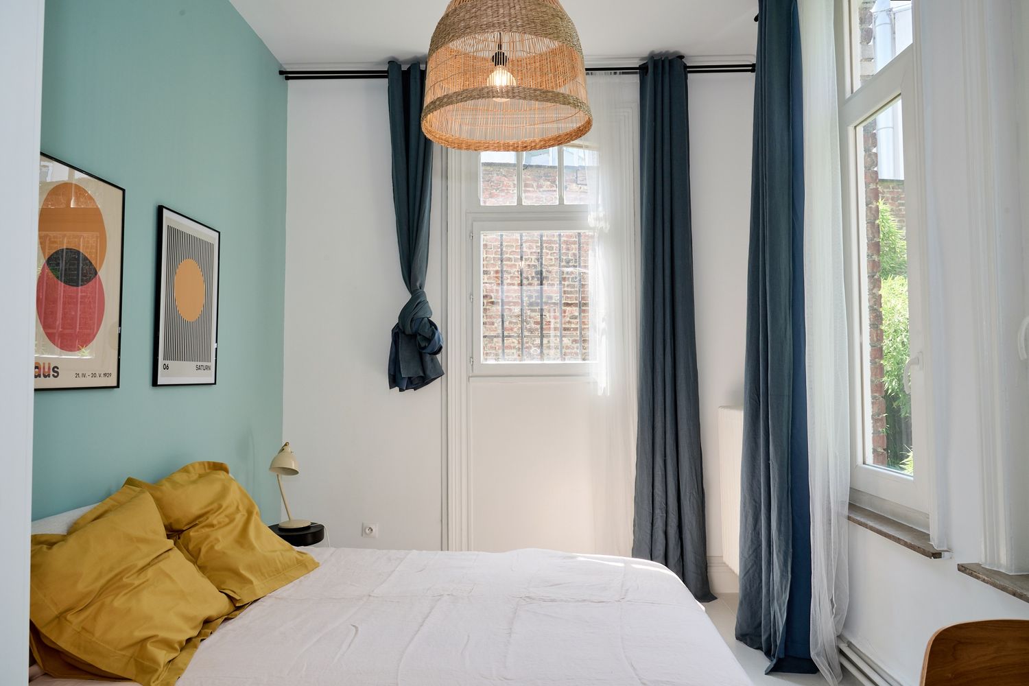 Cozy bedroom with teal walls, mustard yellow pillows, white bedding, blue curtains, and a woven pendant light.
