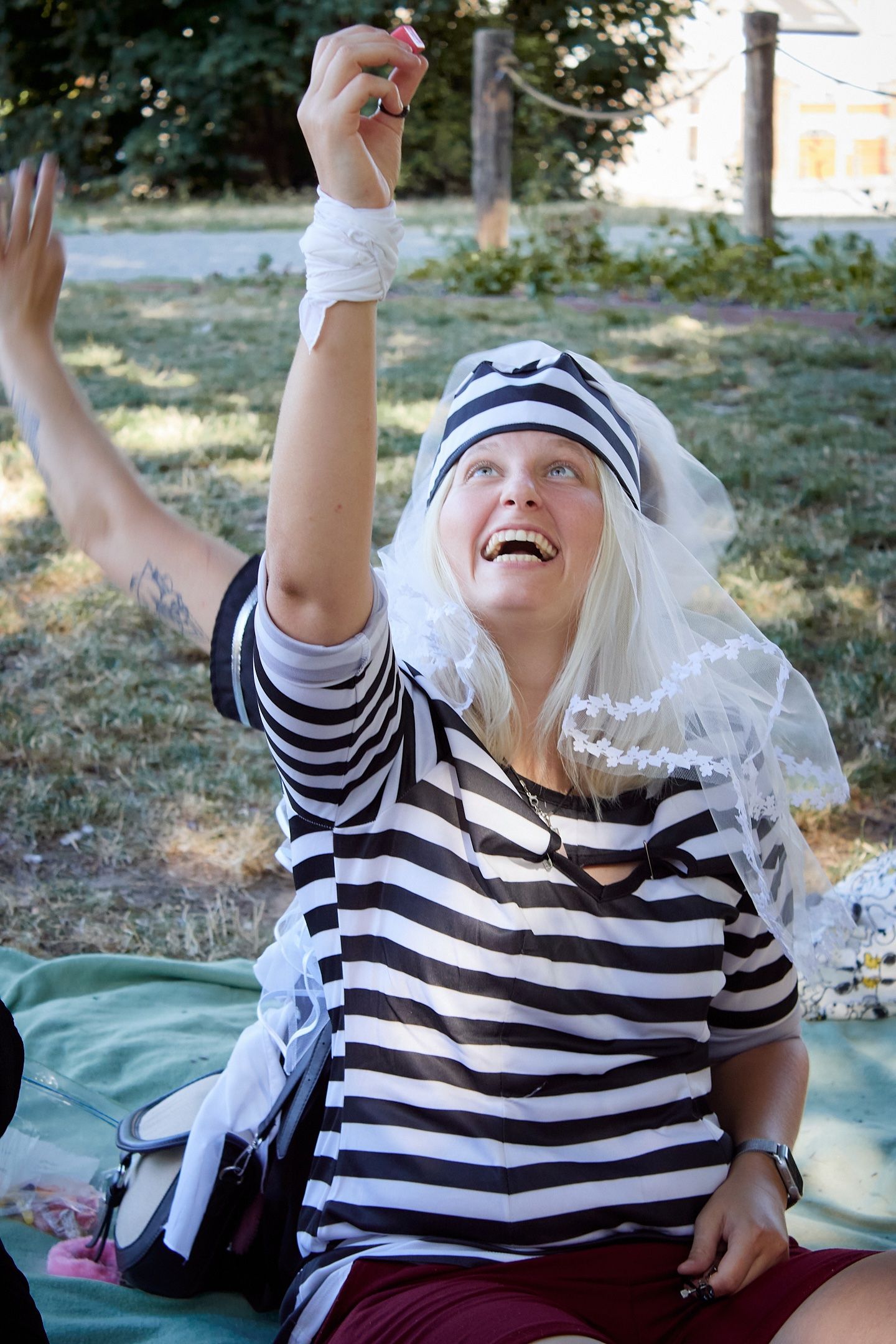 Woman in striped shirt and white wig laughing outdoors, raising her hand in a park setting.