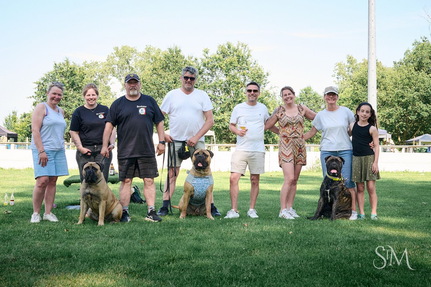 Group of people posing outdoors on grass with their large dogs on a sunny day at a park.
