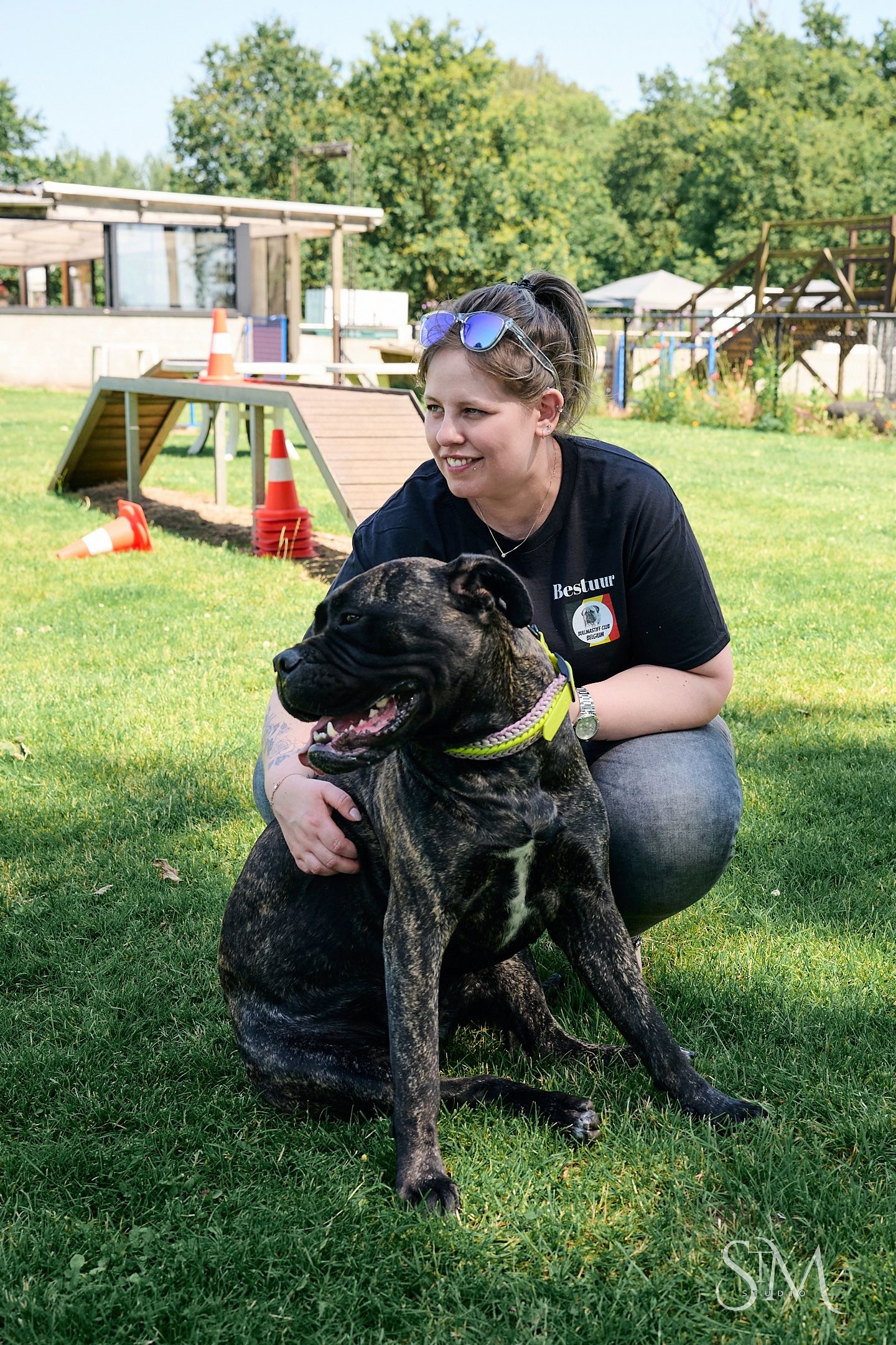 Woman in black shirt crouching beside a brindle dog on green grass at an outdoor dog training facility.