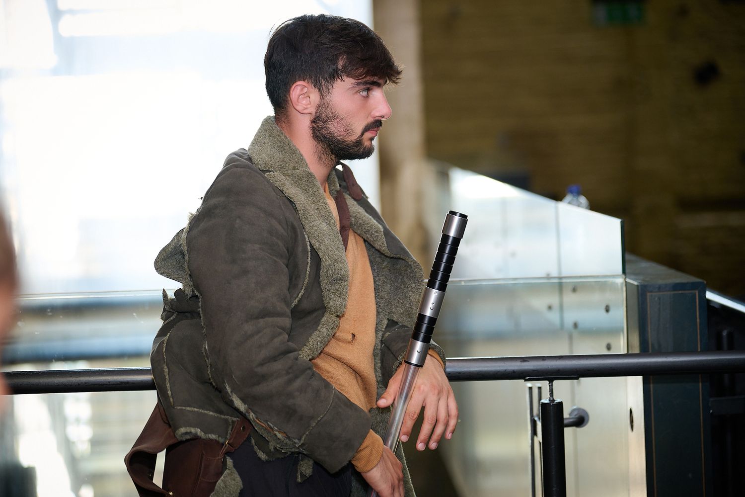 Bearded man in rugged post-apocalyptic costume carrying a weapon and backpack at an indoor event.
