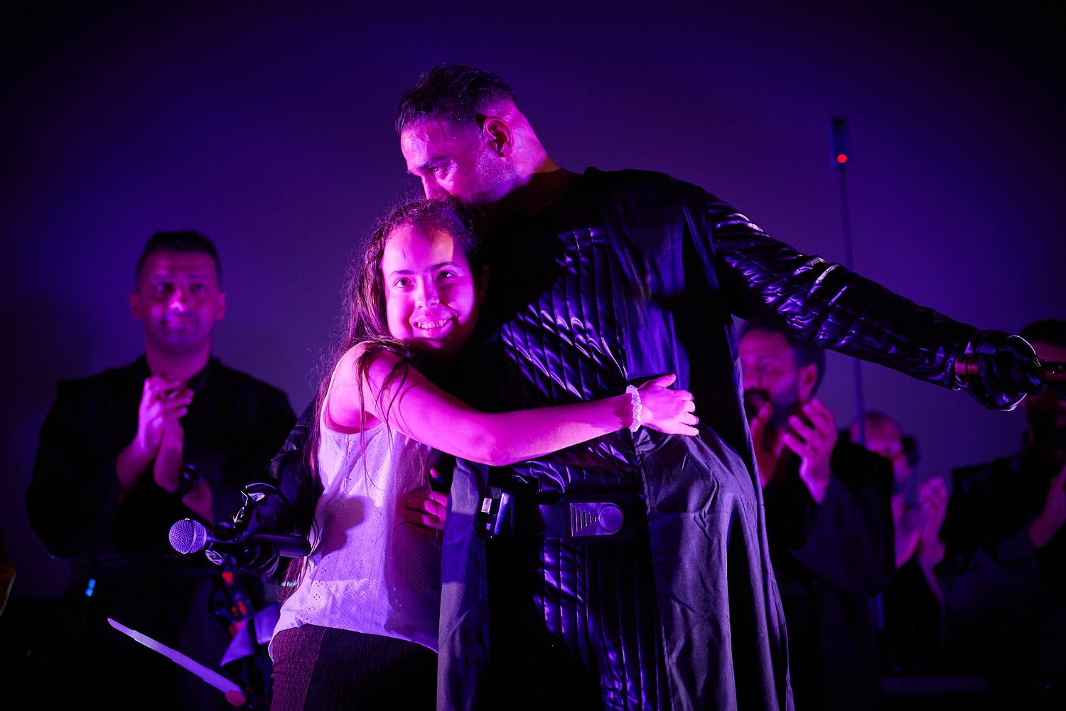 A man in a black jacket hugs a woman on stage under purple lights with musicians in the background.