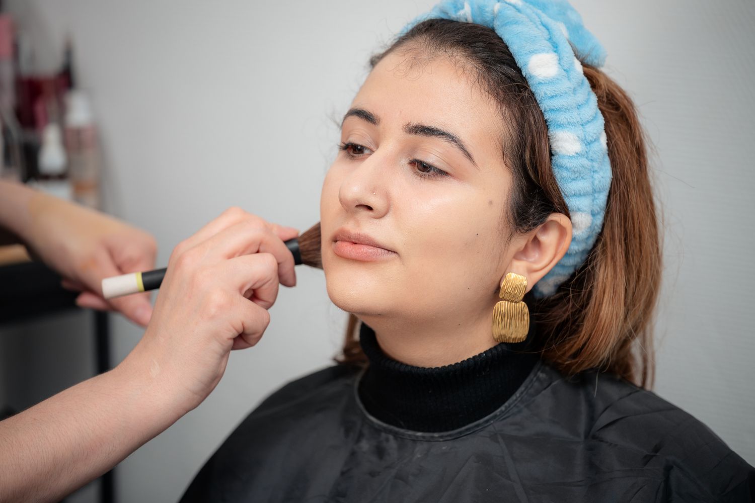 Professional makeup artist applying foundation brush to young woman wearing gold earrings in beauty salon.