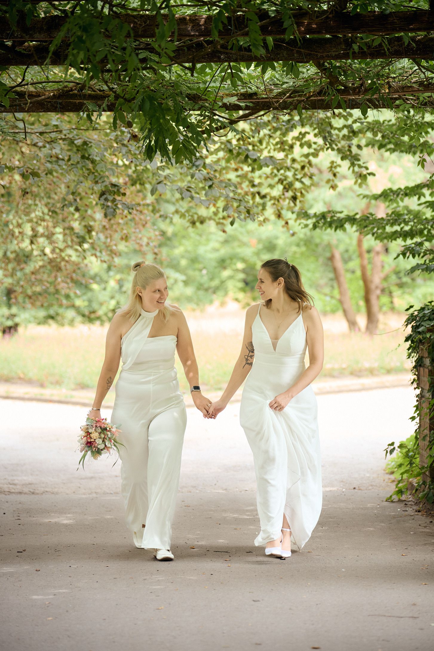 Two brides in white gowns hold hands and walk through a lush green garden pergola on their wedding day.