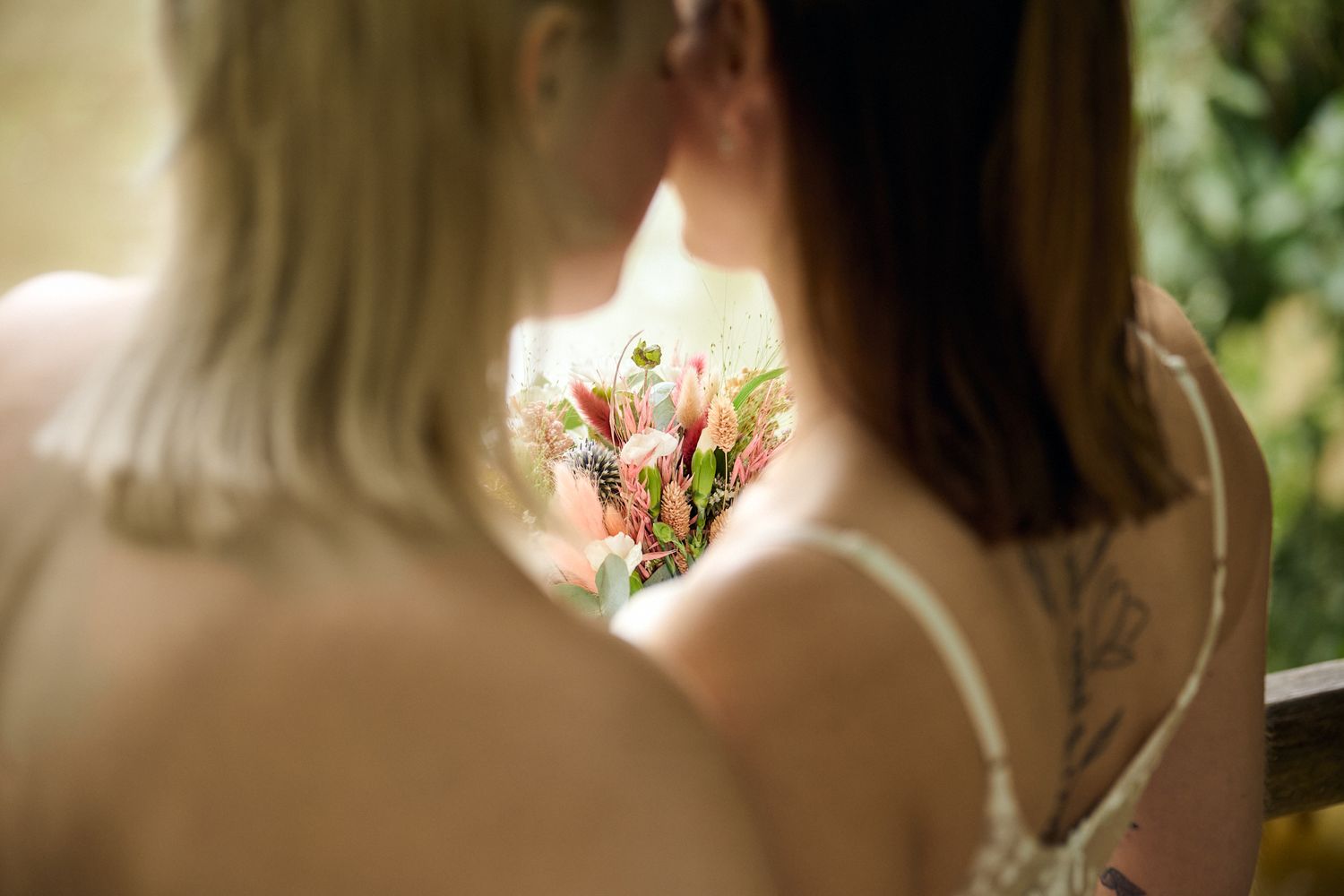 Two brides share an intimate moment, one holding a beautiful pink protea bouquet, with a floral tattoo visible.