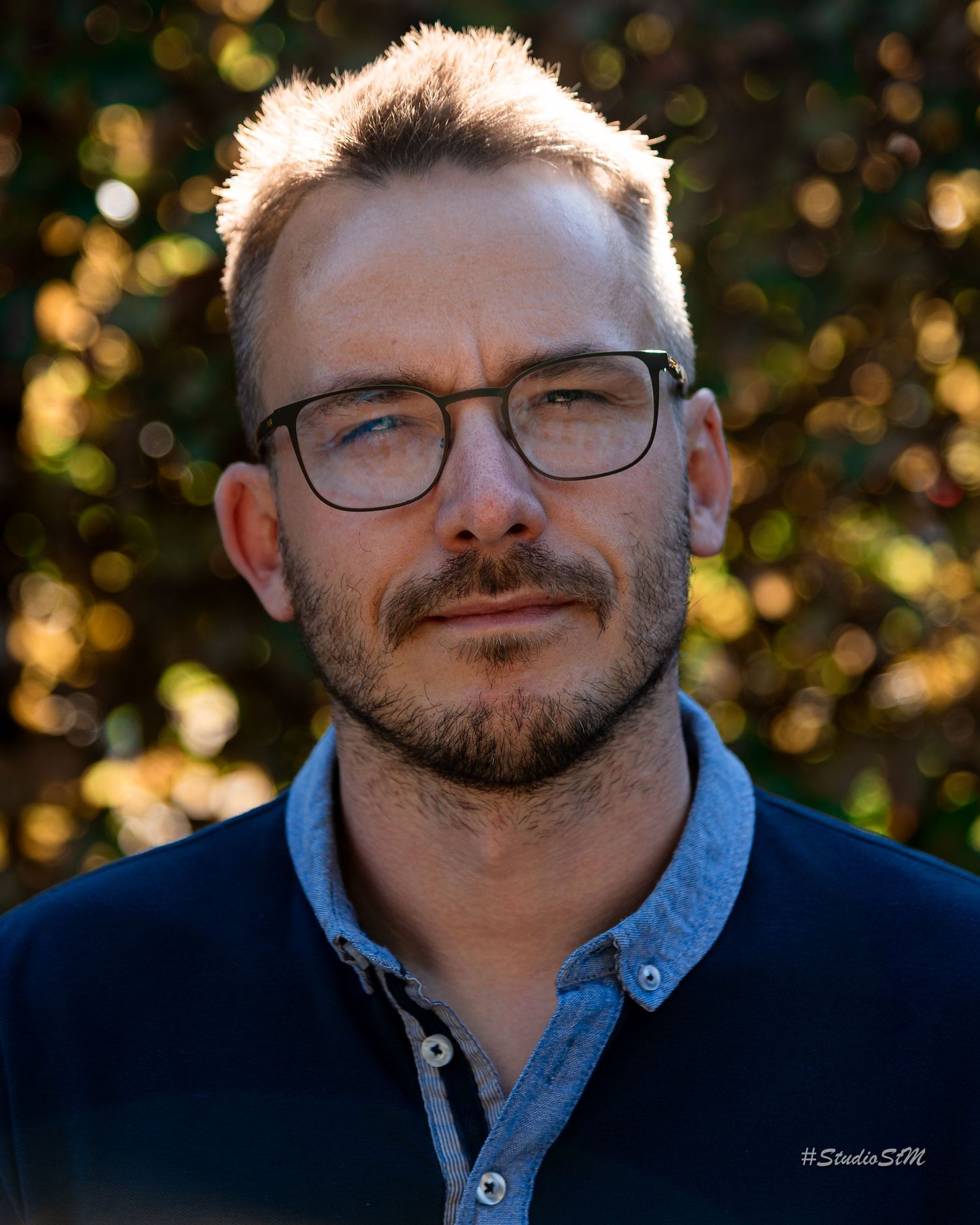 Man with glasses and short hair poses outdoors with bokeh background in professional portrait photo.