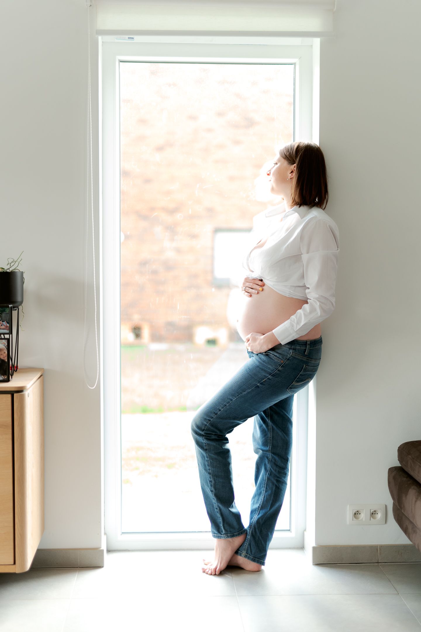 Pregnant woman in white shirt and jeans standing barefoot by a bright window, cradling her baby bump at home.