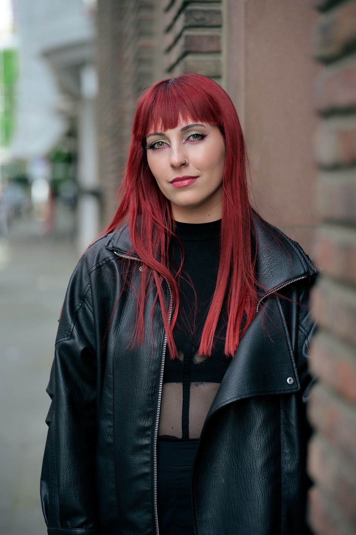 Woman with long red hair in black leather jacket posing against brick wall outdoors.