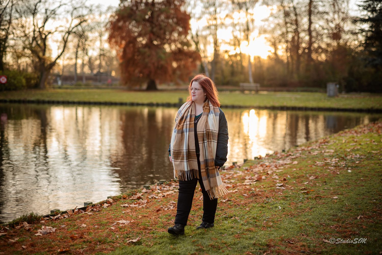 Red-haired woman in plaid scarf standing by reflective pond in autumn park at golden hour.
