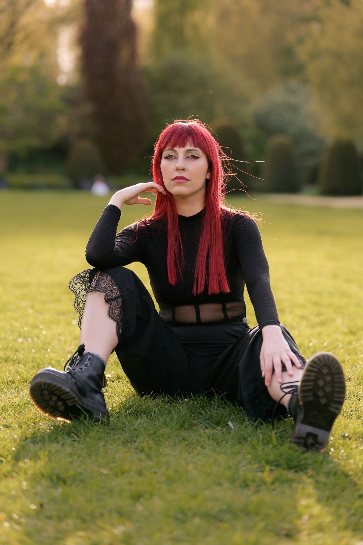 Red-haired woman in black outfit sitting on grass in sunny park with boots.