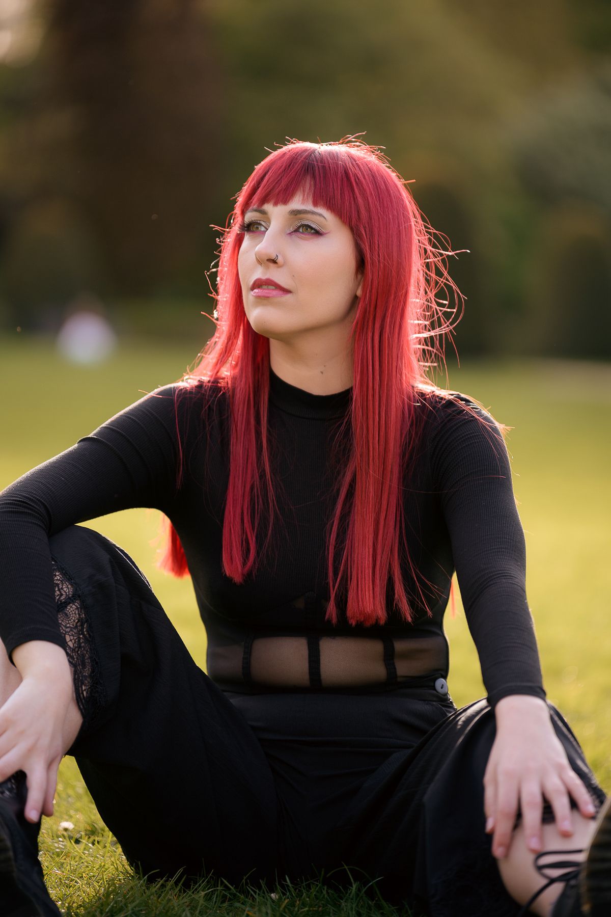 A woman with vibrant red hair sits on green grass wearing an all-black outfit, gazing thoughtfully in a sunlit park.