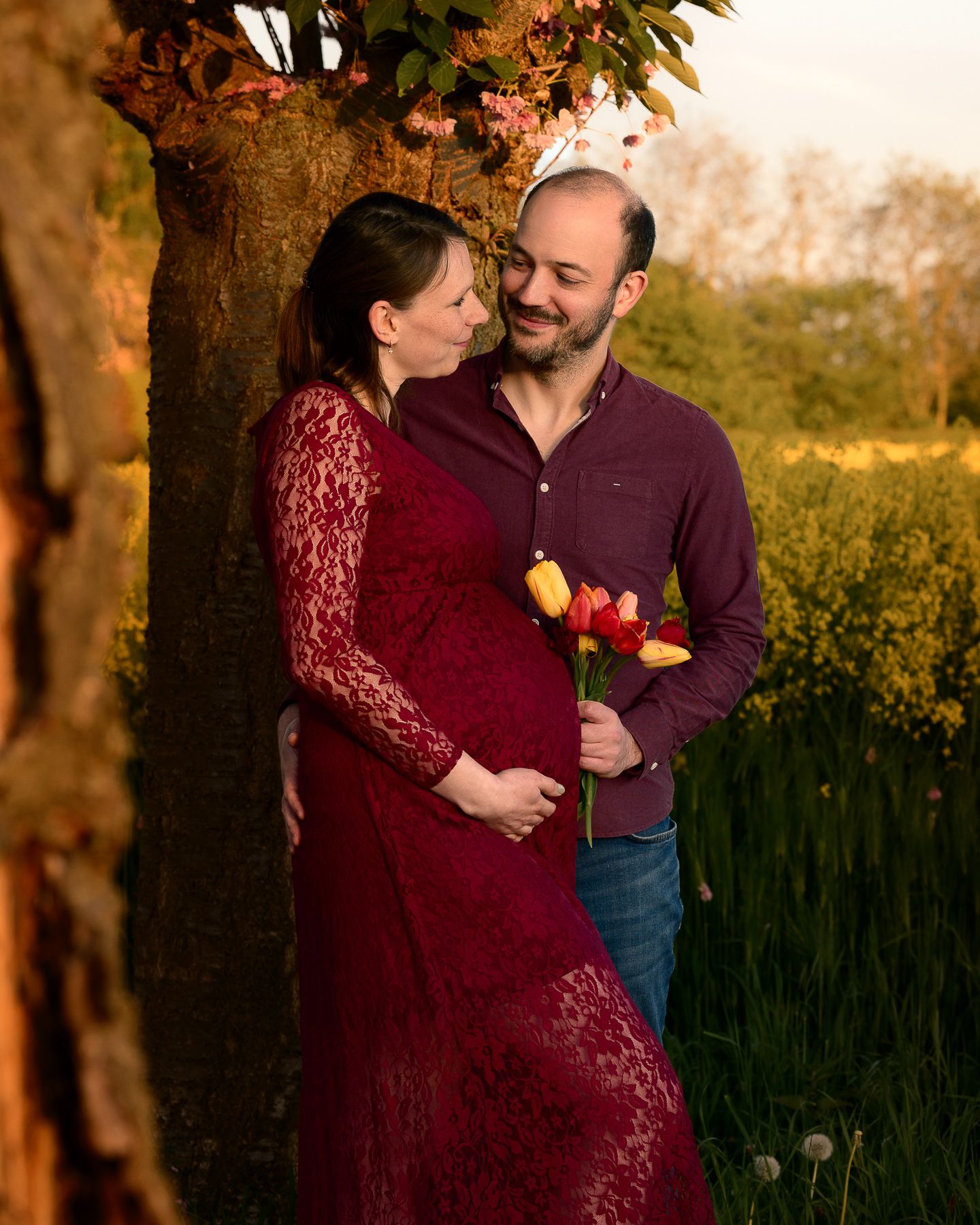 Couple sharing tender moment outdoors at golden hour, woman in red lace maternity gown holding tulips with partner.