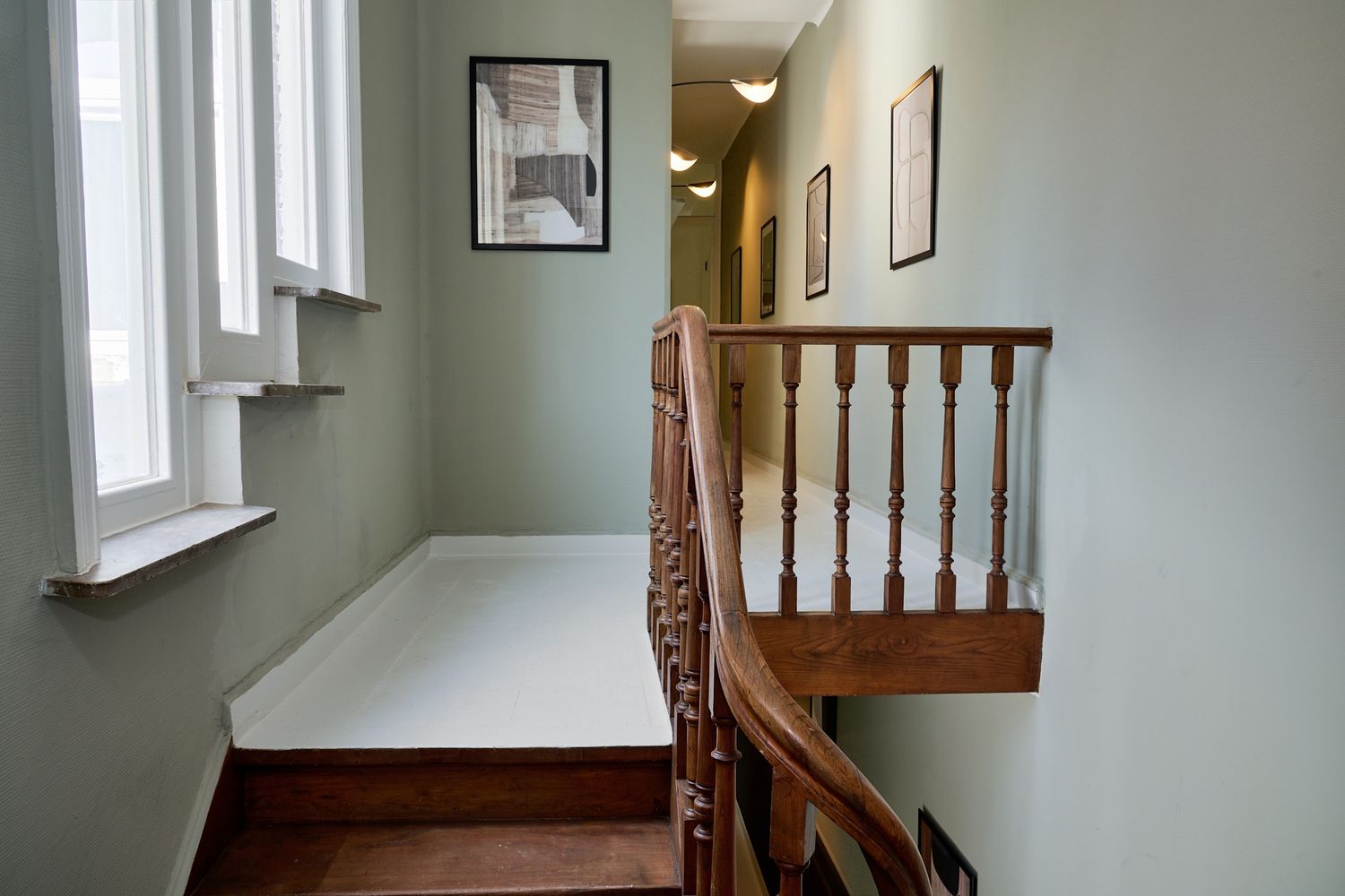 Elegant hallway with wooden staircase railing, sage green walls, framed artwork, and white window seat in a classic home.