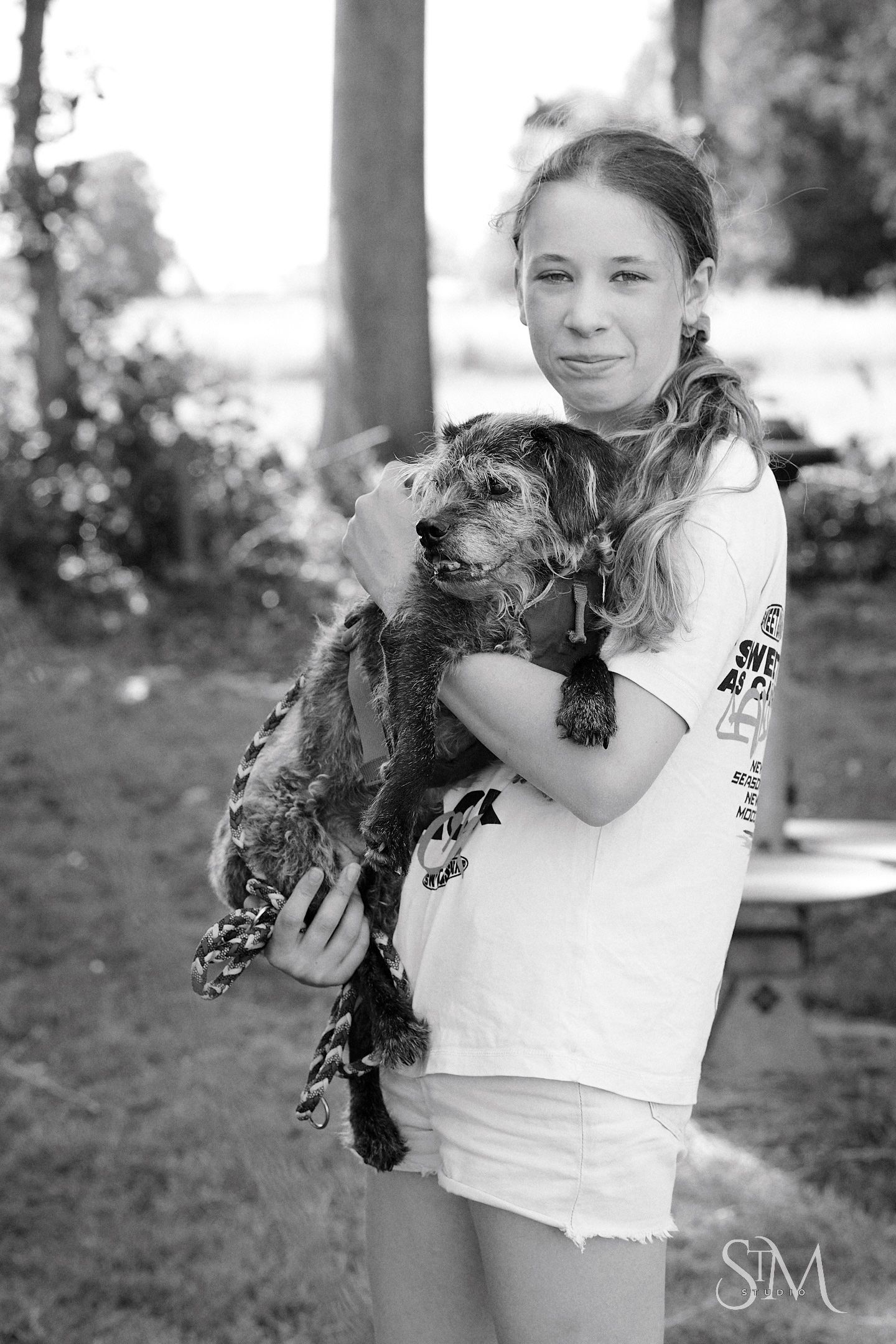 Black and white photo of a young girl holding a small scruffy dog outdoors near trees.