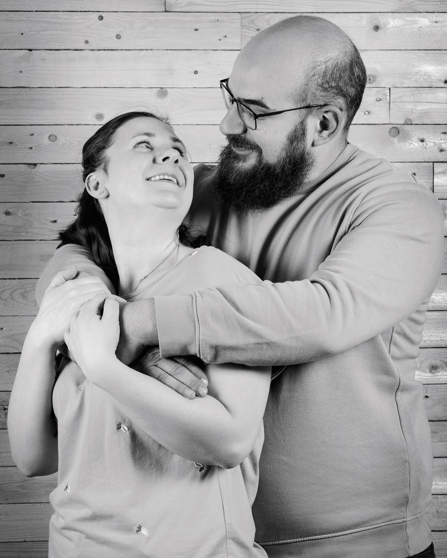 Black and white photo of a smiling couple embracing, bald bearded man with glasses hugging a laughing woman against wood wall.