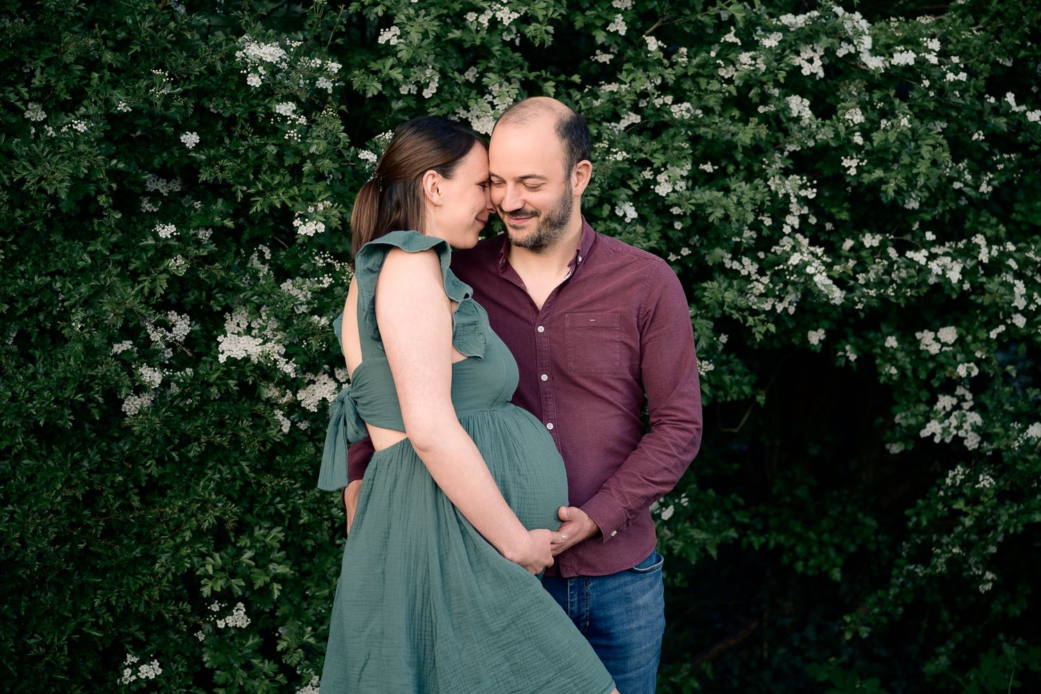 Couple sharing tender moment outdoors, pregnant woman in green dress with partner, surrounded by white flowering bushes.