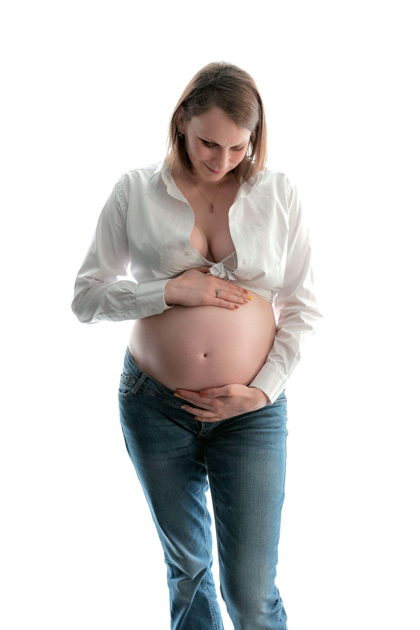 Pregnant woman in white shirt and jeans cradling her bare baby bump, smiling downward on white background.