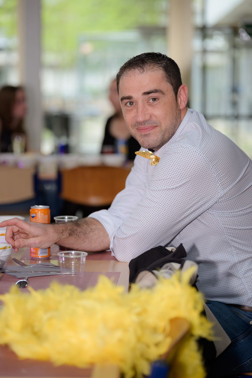 Man in white shirt smiling at table with yellow decorations and drinks at an indoor event.