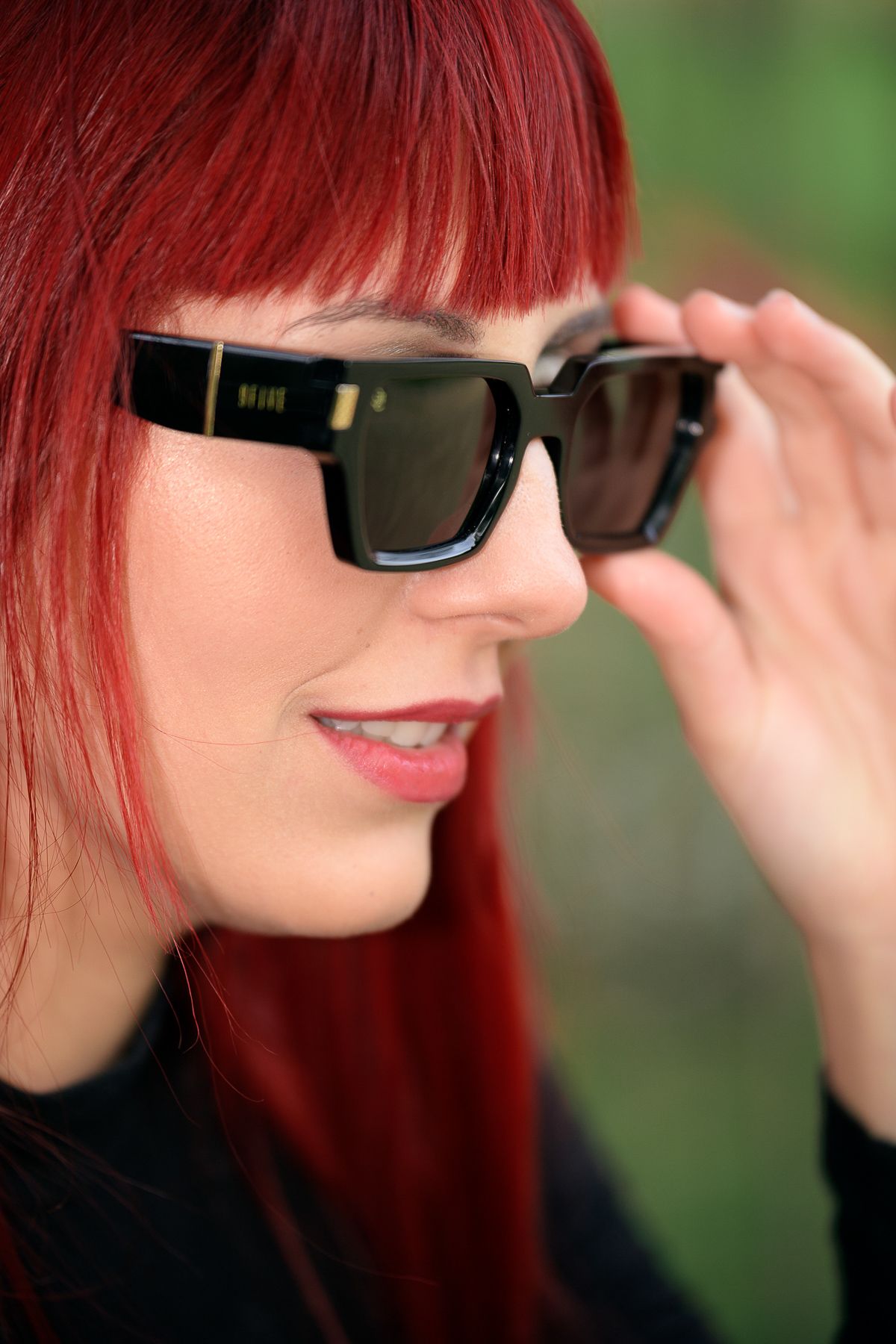 Red-haired woman adjusting black square sunglasses outdoors, smiling slightly against a blurred green background.