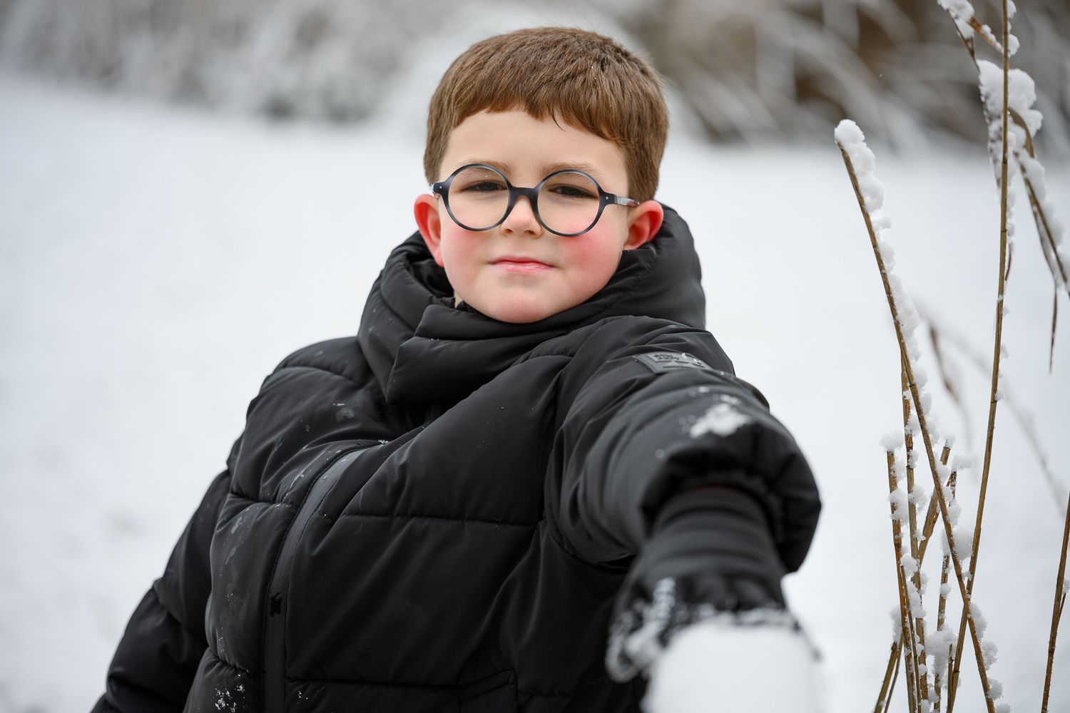 A young boy wearing glasses and a black winter jacket stands in a snowy outdoor setting.