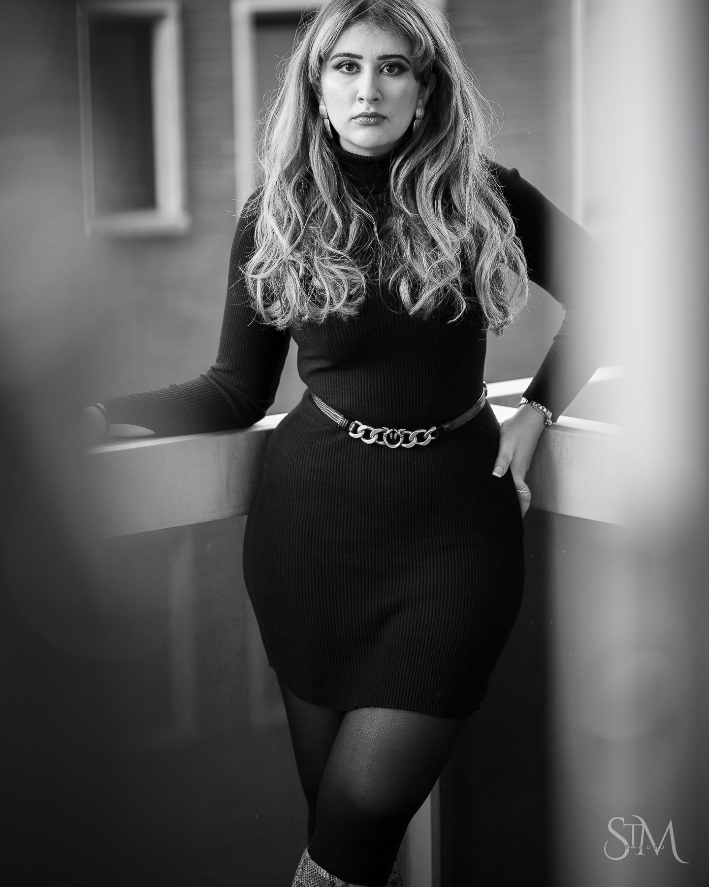 Woman in black turtleneck dress and chain belt poses confidently in a stylish black and white portrait photo.