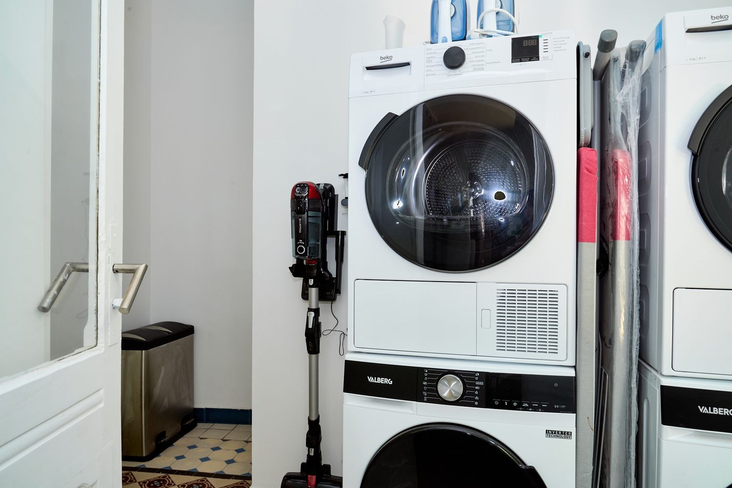 Stacked white washer and dryer units in a clean laundry room with a vacuum cleaner beside them.