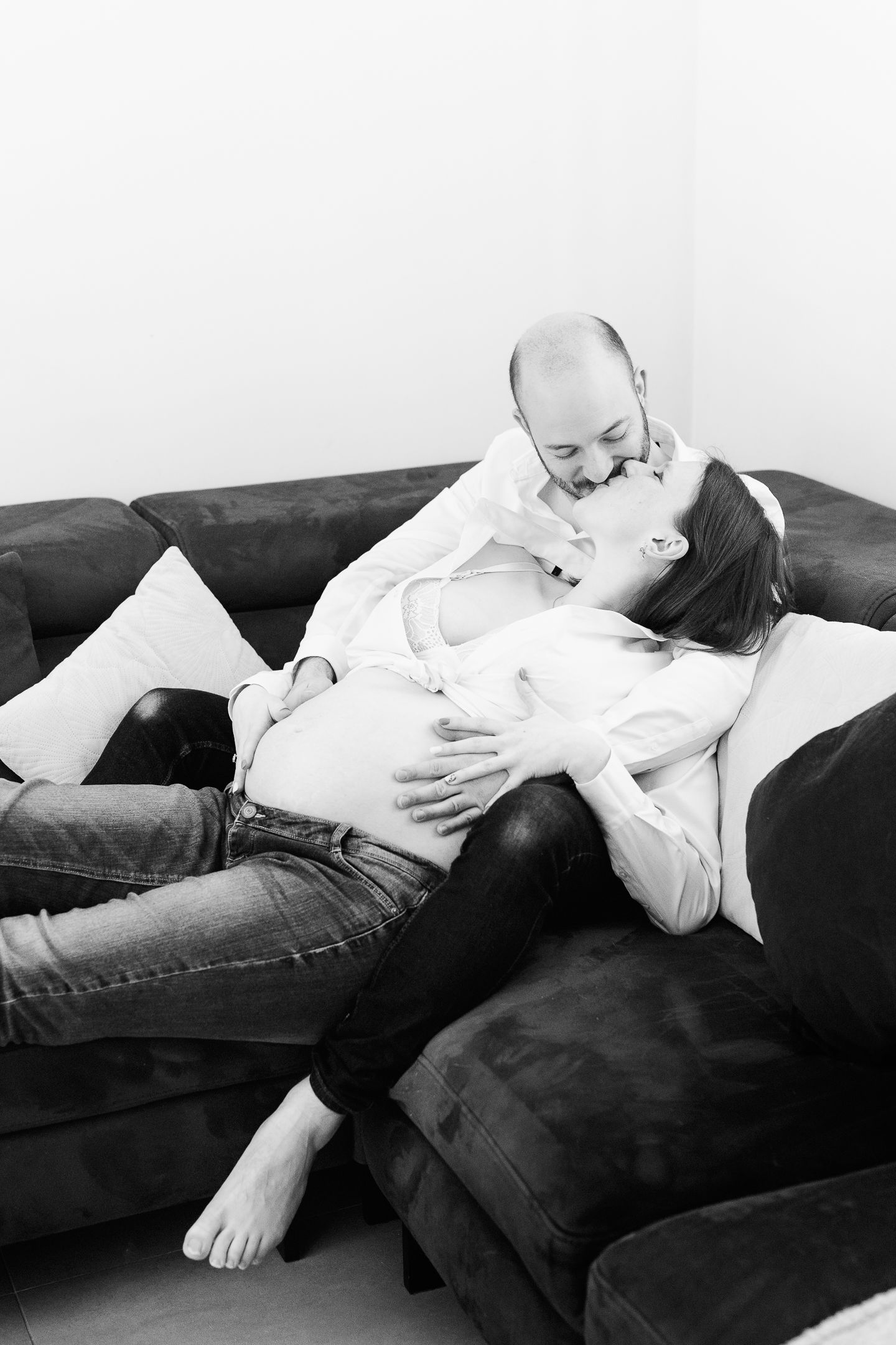 Black and white photo of a pregnant couple kissing on a dark sofa, woman lying with baby bump visible.
