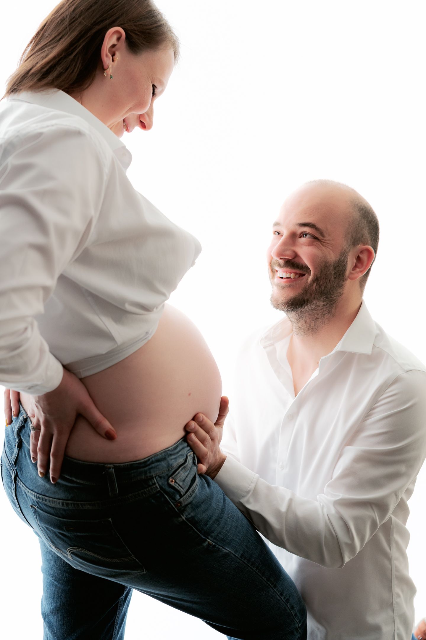 Happy couple expecting a baby, man smiling at pregnant woman's bare belly in a bright studio setting.