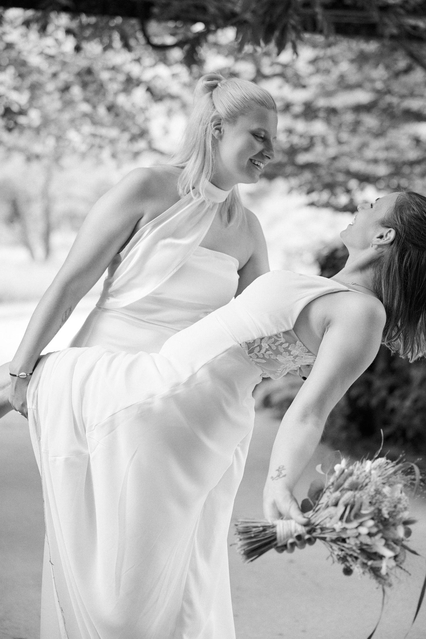 Black and white photo of two brides in white gowns laughing joyfully, one holding a bouquet outdoors.