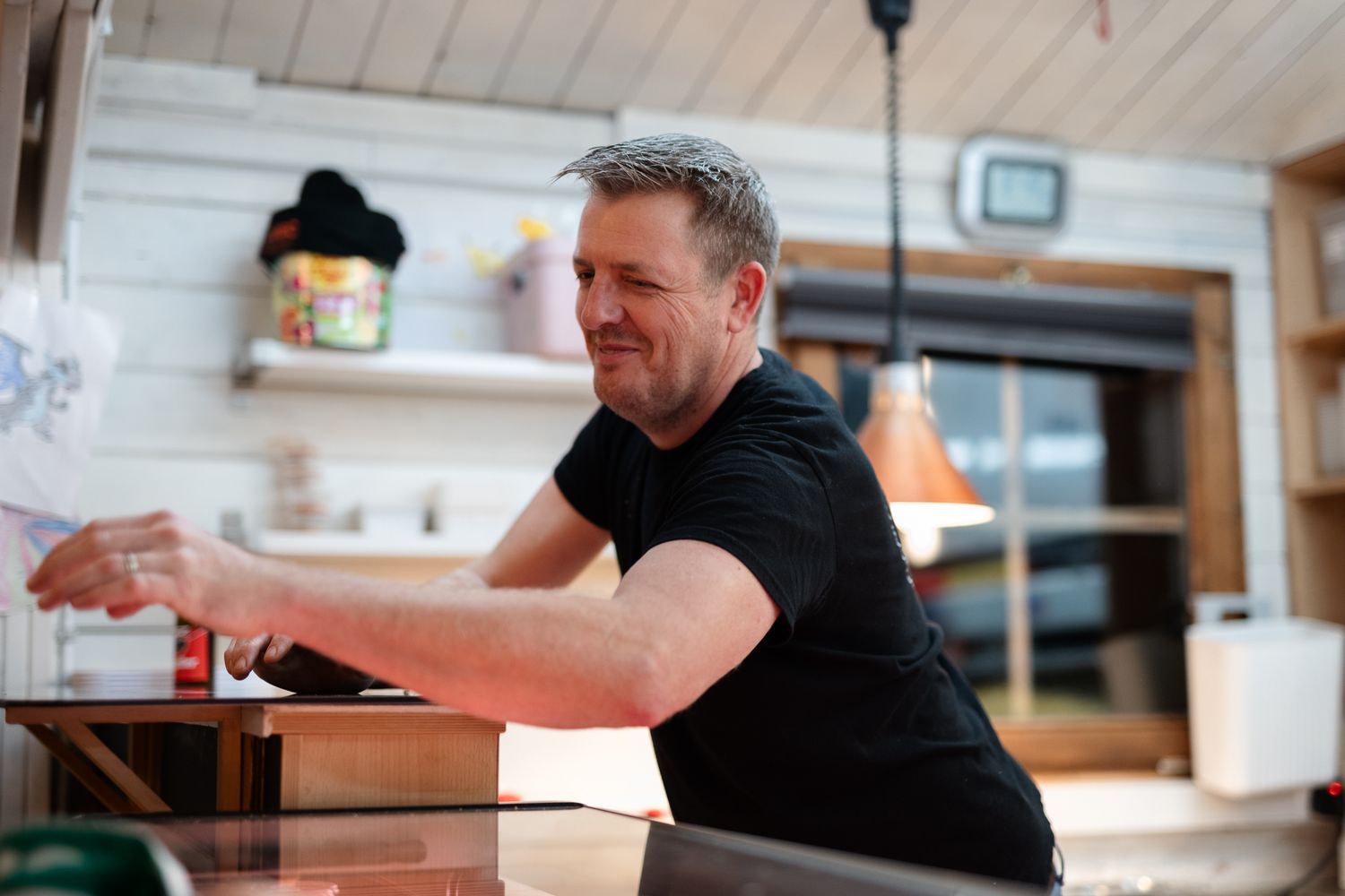 Man in black t-shirt leaning over a wooden counter in a cozy kitchen interior.