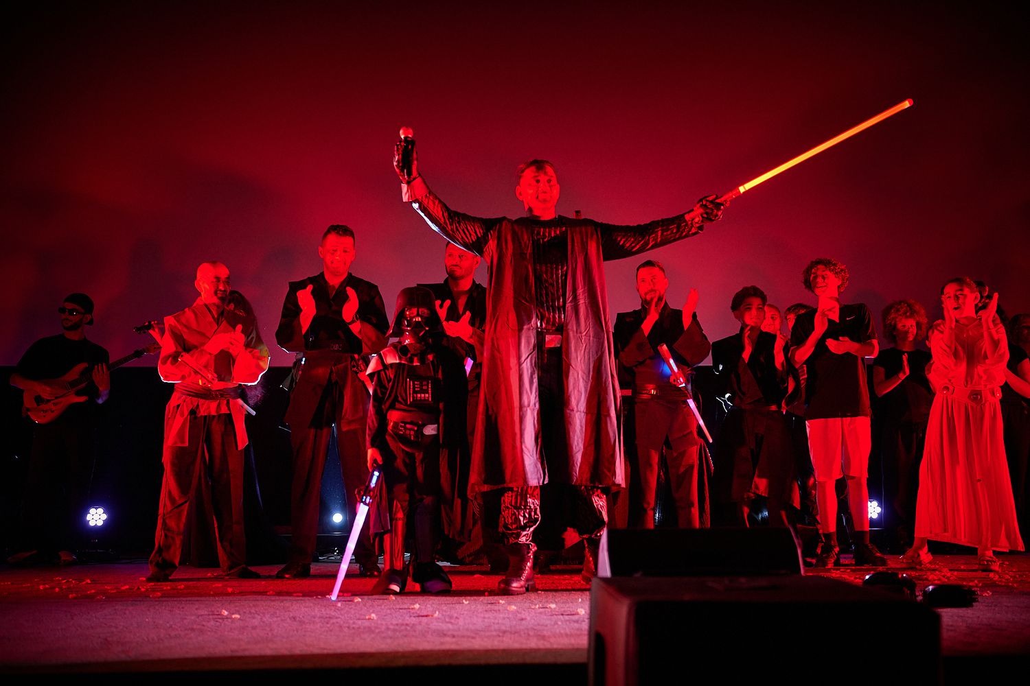 Star Wars Sith cosplayer raises lightsabers triumphantly on a dramatic red-lit performance stage.