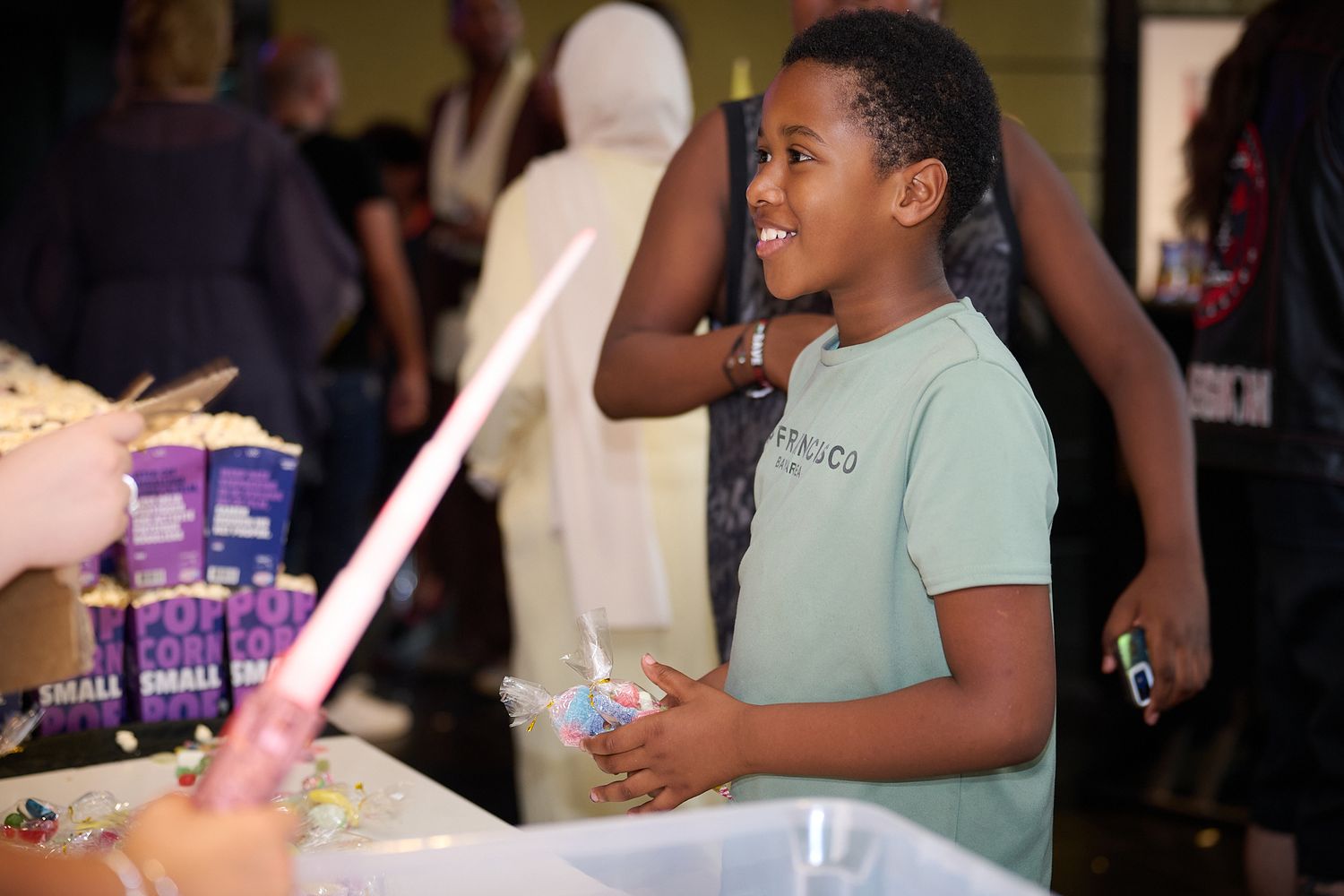 A young boy smiles at a community event table with snacks and activities in a busy indoor venue.