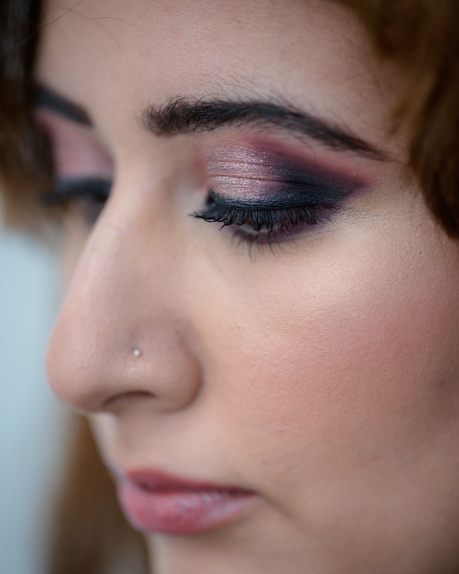 Close-up of a woman wearing dramatic smoky eye makeup with dark burgundy and pink eyeshadow and a nose stud.
