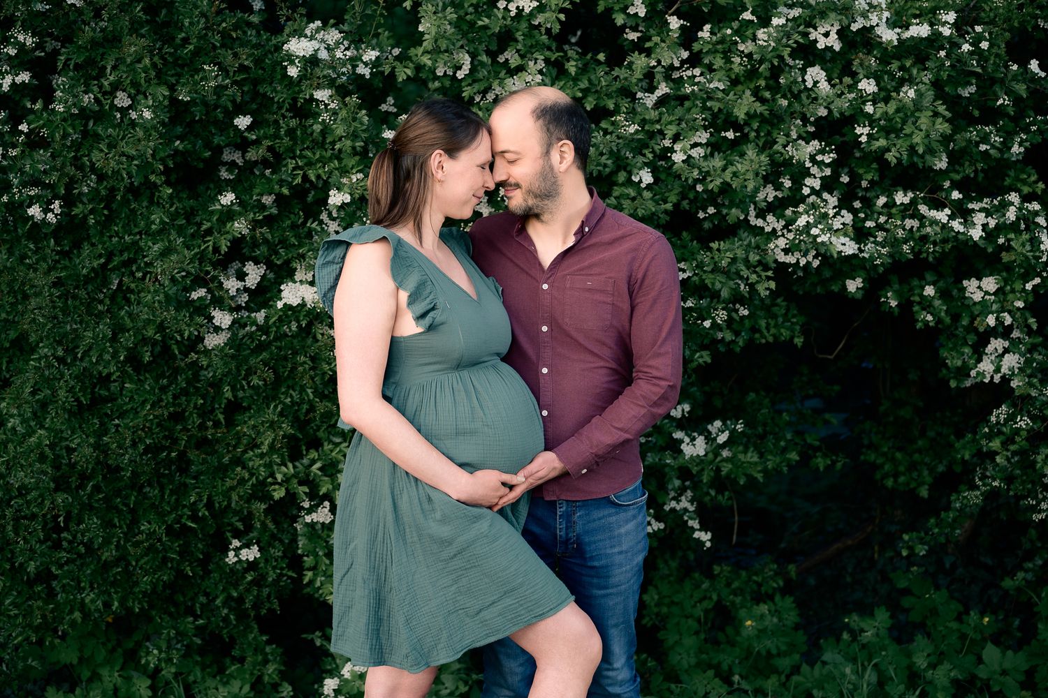 Pregnant couple embracing outdoors in front of lush green flowering bushes, woman in green dress, man in burgundy shirt.