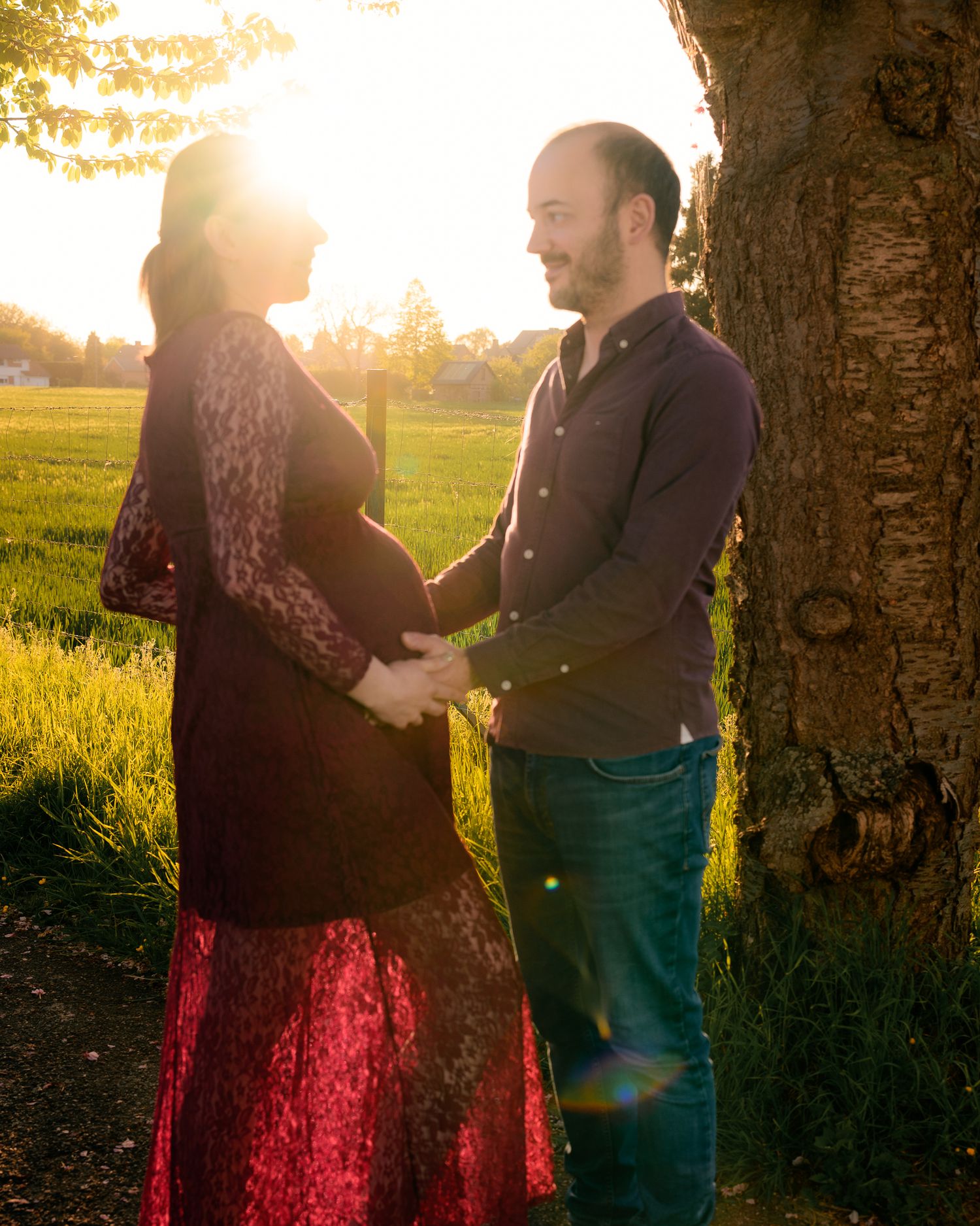 Couple sharing a tender moment outdoors at golden hour, woman in red lace maternity dress, man in navy shirt holding her bump.