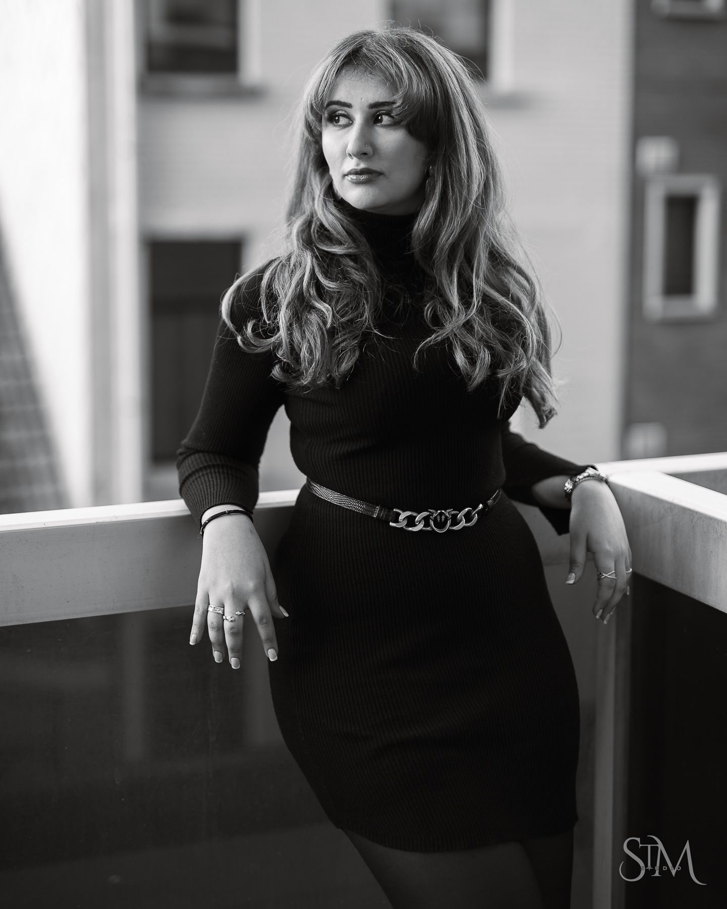 Elegant black and white portrait of a woman in a black dress with chain belt leaning on a railing.