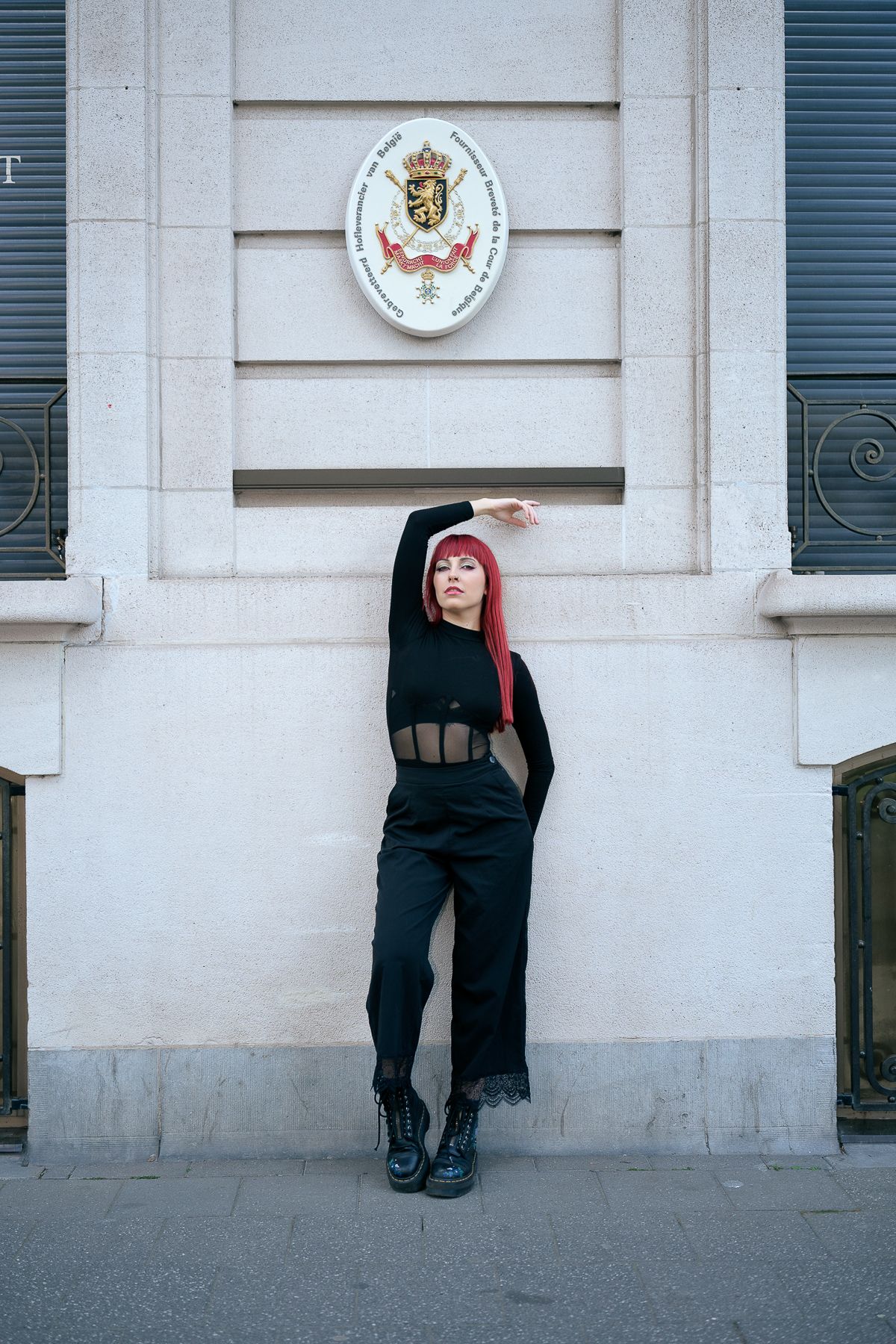 Red-haired woman in black outfit posing against a European building with a coat of arms.