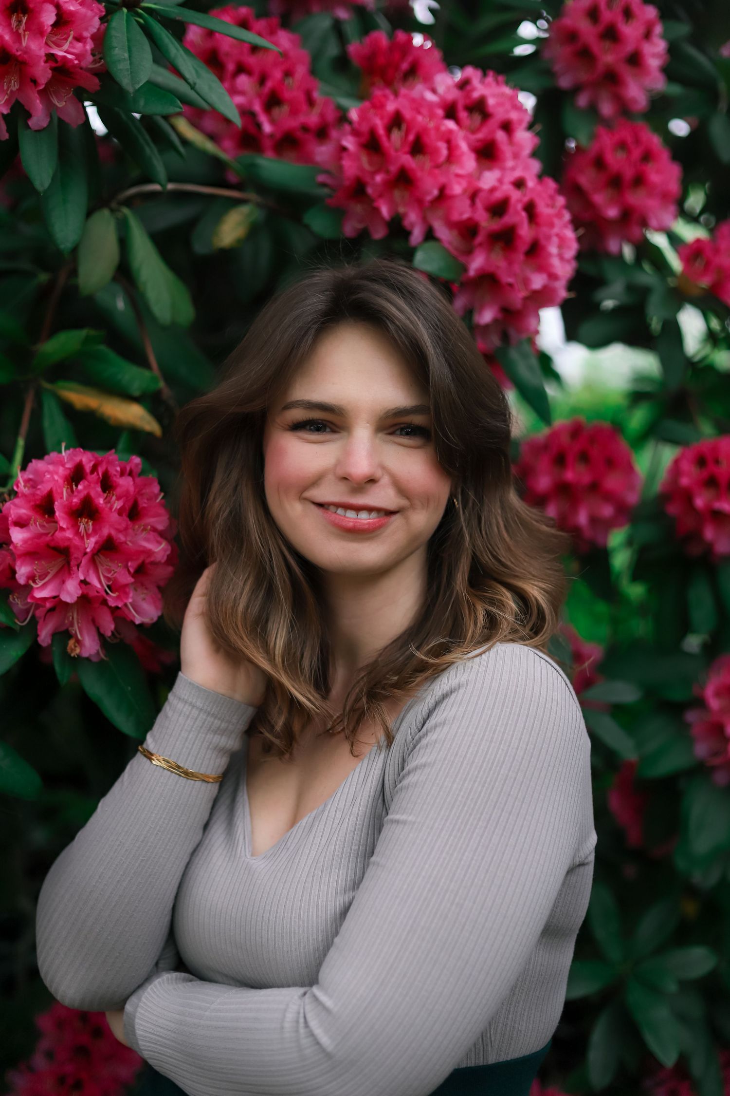 Beautiful young woman with wavy brown hair smiling warmly against a backdrop of pink blooms.