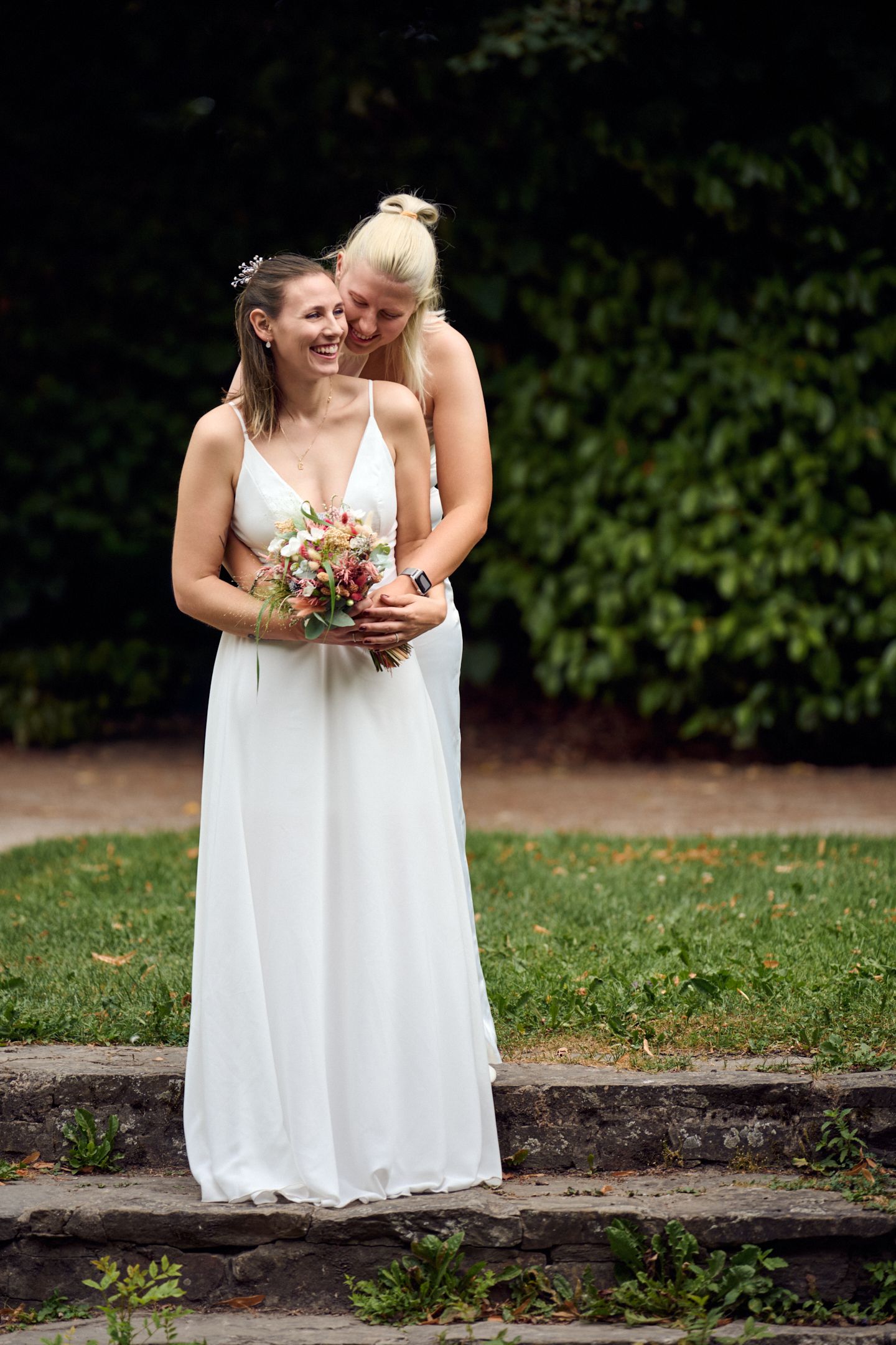 Two brides in white dresses embrace outdoors, one holding a colorful bouquet, smiling joyfully on stone steps.