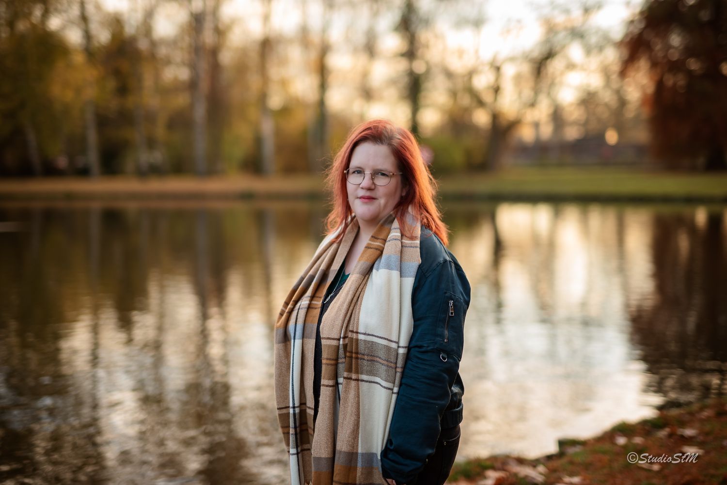 Red-haired woman wearing plaid scarf standing near pond surrounded by autumn foliage in park.