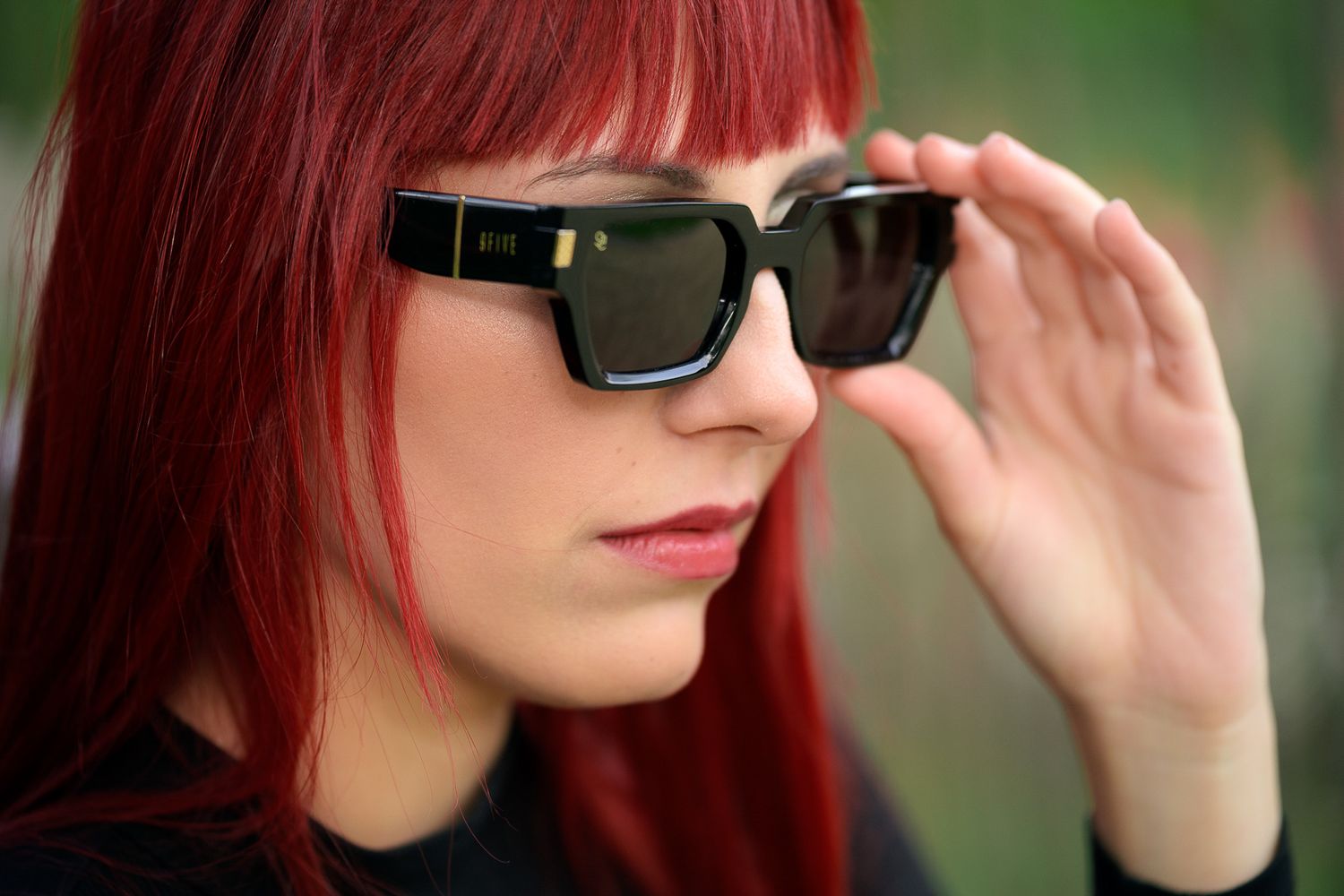 Young woman with vibrant red hair adjusting black square sunglasses outdoors.